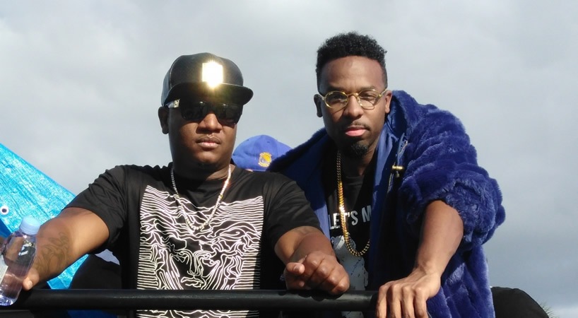 Two men standing outdoors, one wearing sunglasses and a cap, the other with glasses and a blue jacket.