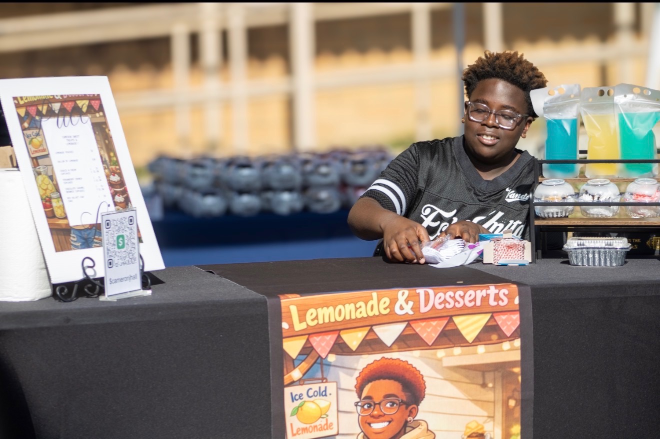 Young person smiling behind lemonade and dessert stand with colorful sign and drinks in background.