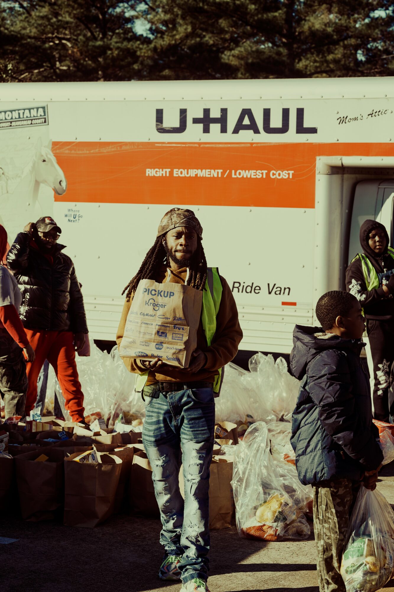 Person with dreadlocks holding a bag, standing outdoors near a U-Haul truck and other people.