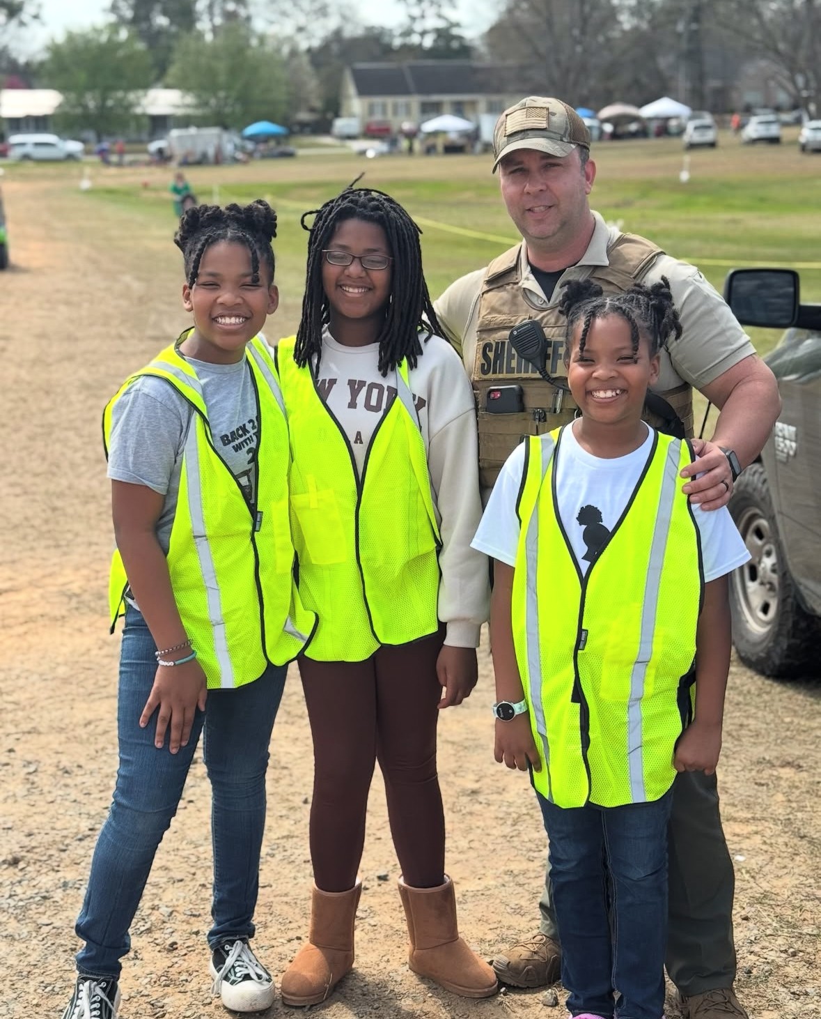 Three young girls and a man in uniform stand outdoors on a grassy field, smiling at the camera.