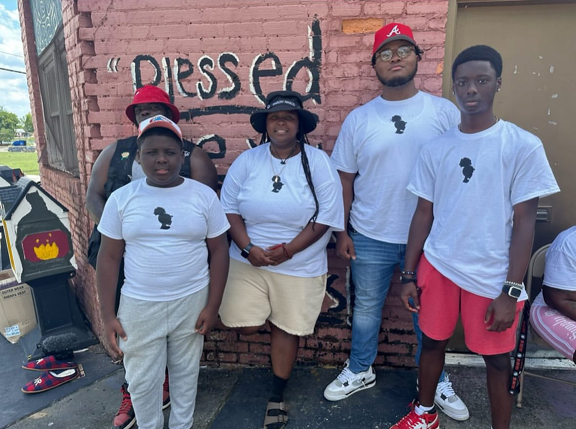 Four people stand in front of a brick wall with graffiti, wearing white shirts with black silhouette logos.