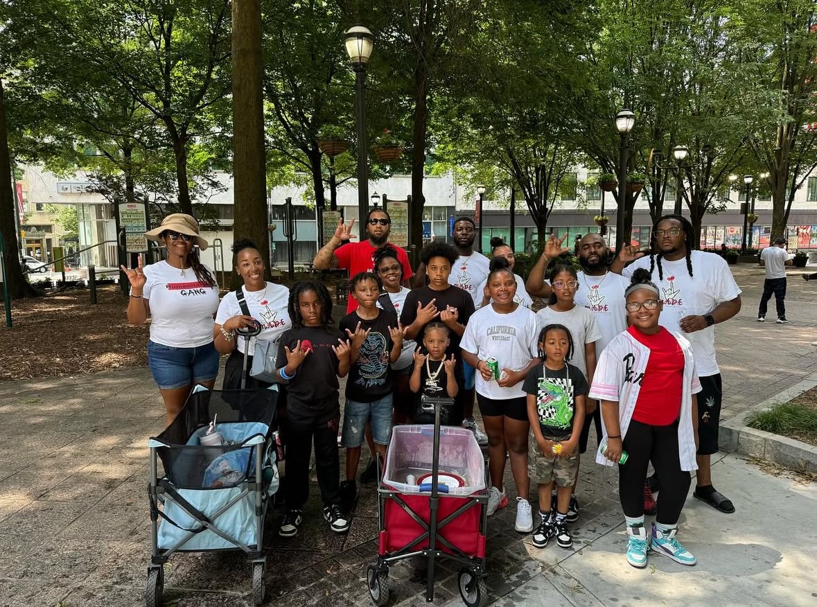 Group of children and adults outdoors in a park with trees and lampposts, some pushing strollers, smiling at the camera.