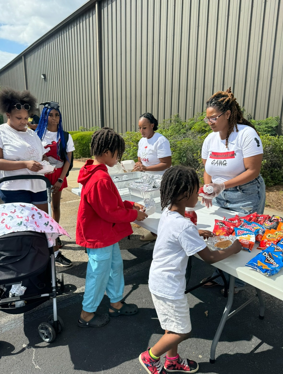 Group of children and a woman at an outdoor table with snacks, near a large metal building and greenery.