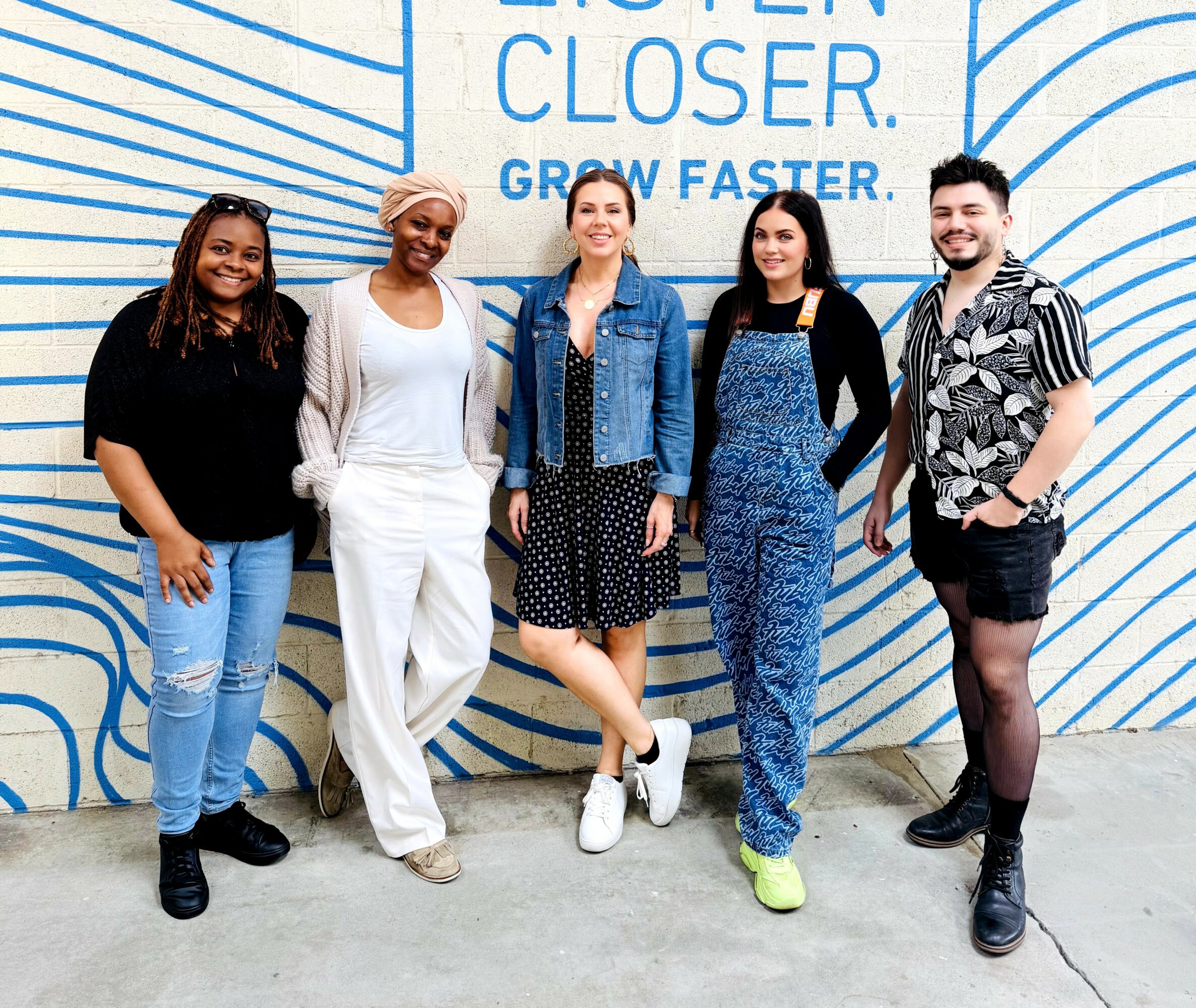Five diverse women standing in front of a blue and white mural with text, smiling at the camera.