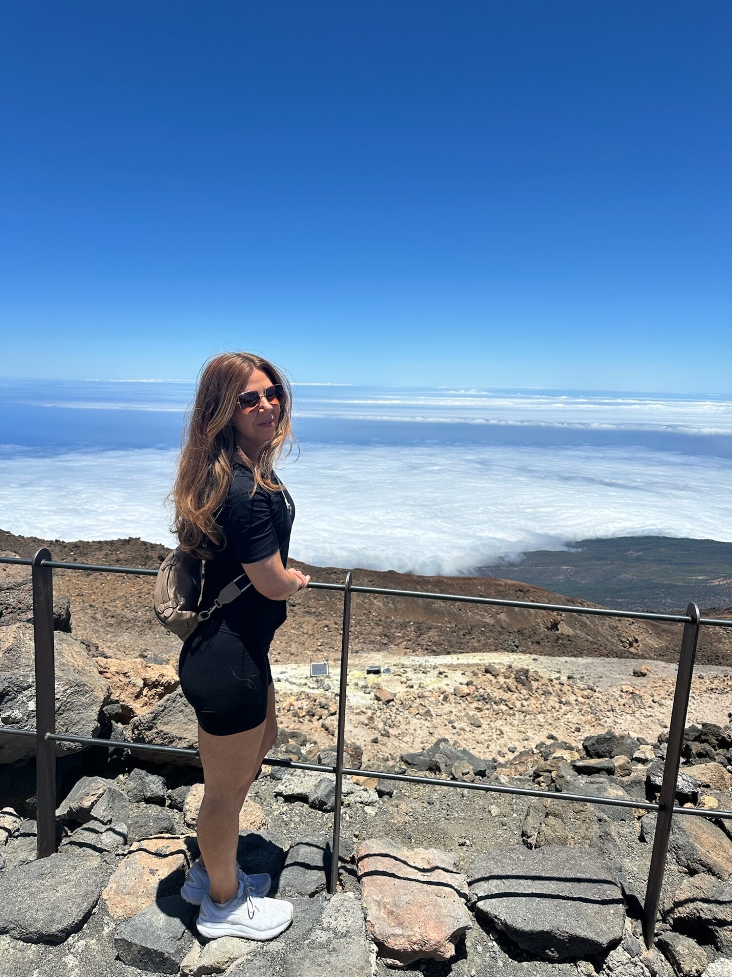Woman with long hair wearing black outfit and white shoes standing by a railing near rocky terrain with a vast salt flat and blue sky in the background.