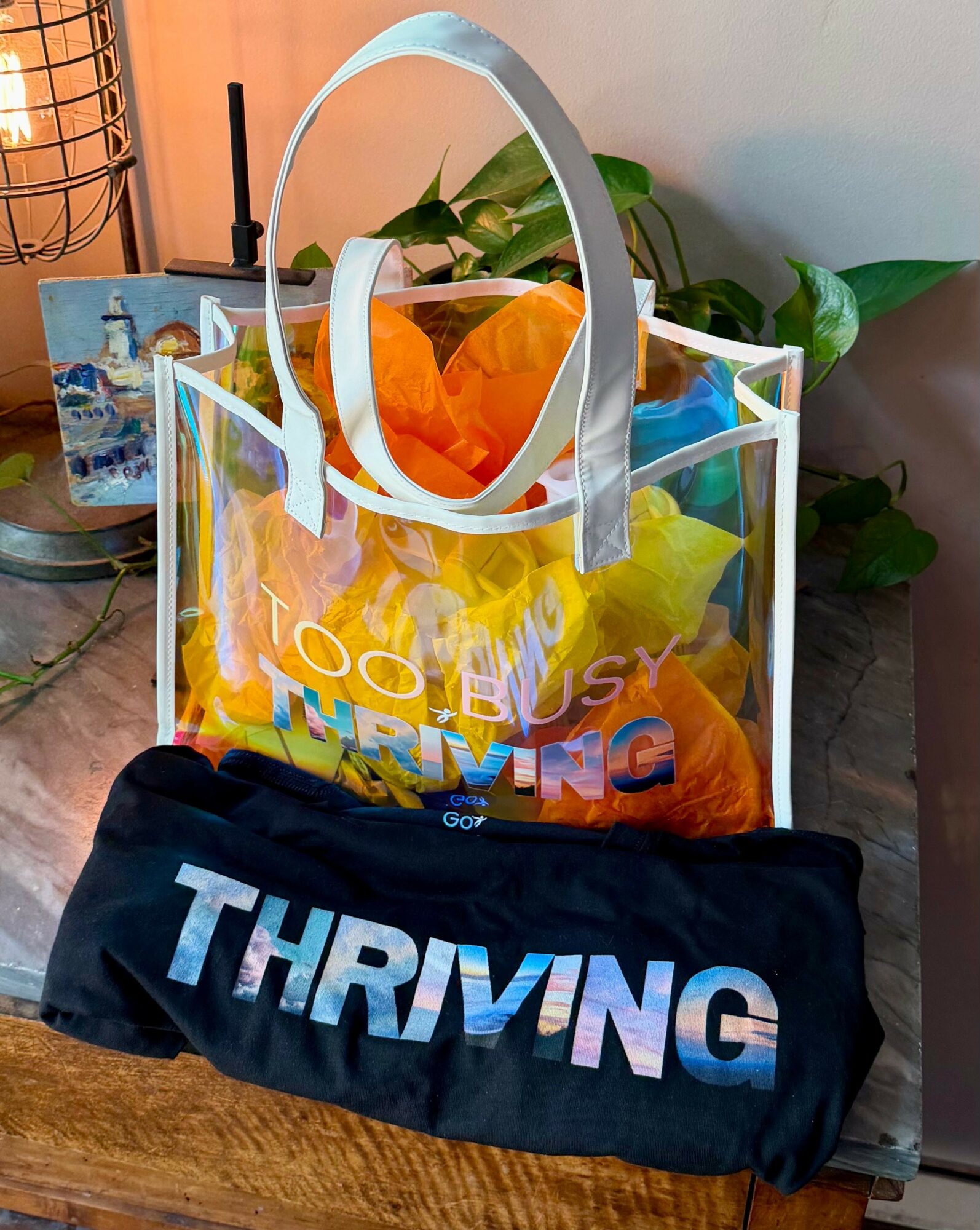 Transparent tote bag with yellow and orange flowers, and colorful text, placed on a wooden surface with a black fabric item in front.