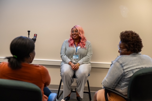 Woman with pink hair speaking to two seated women in a room with a plain wall.