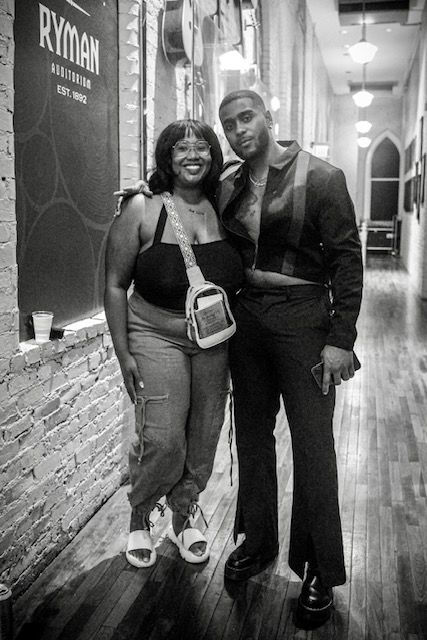 Two people standing together in a hallway, smiling, with a brick wall and hanging lights in the background.