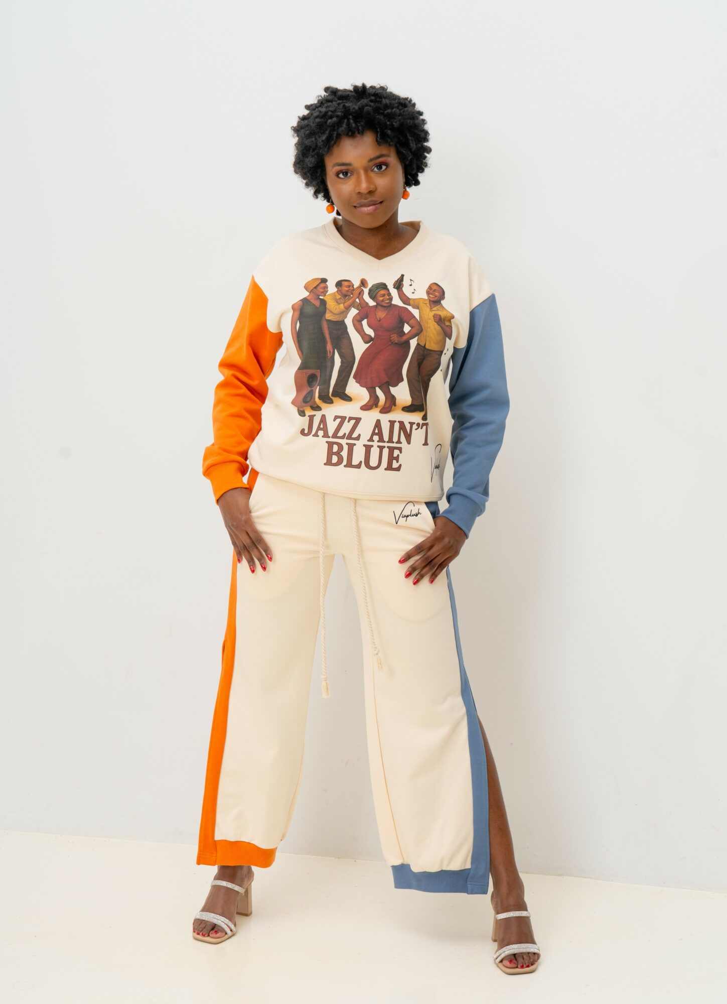 Person with curly hair wearing a colorful patchwork shirt and wide-leg pants standing against a white background.