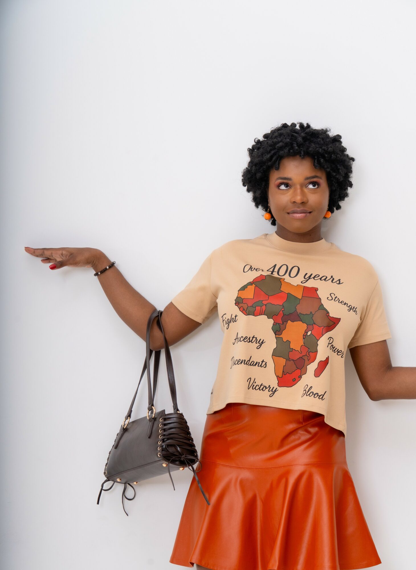 Young woman with curly hair wearing a beige T-shirt and orange skirt, standing against a white background with arms outstretched.