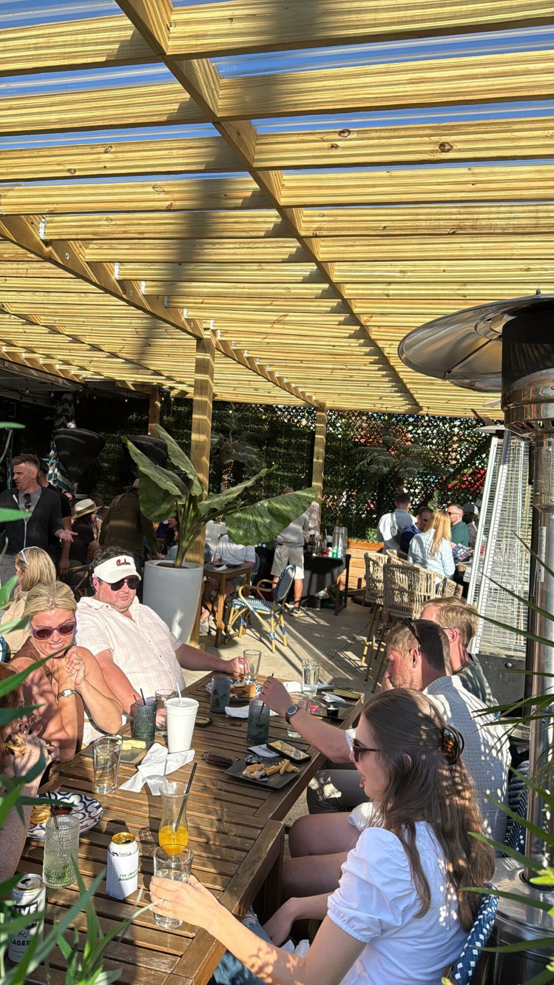 People sitting at an outdoor table under a wooden pergola, enjoying drinks and conversation.