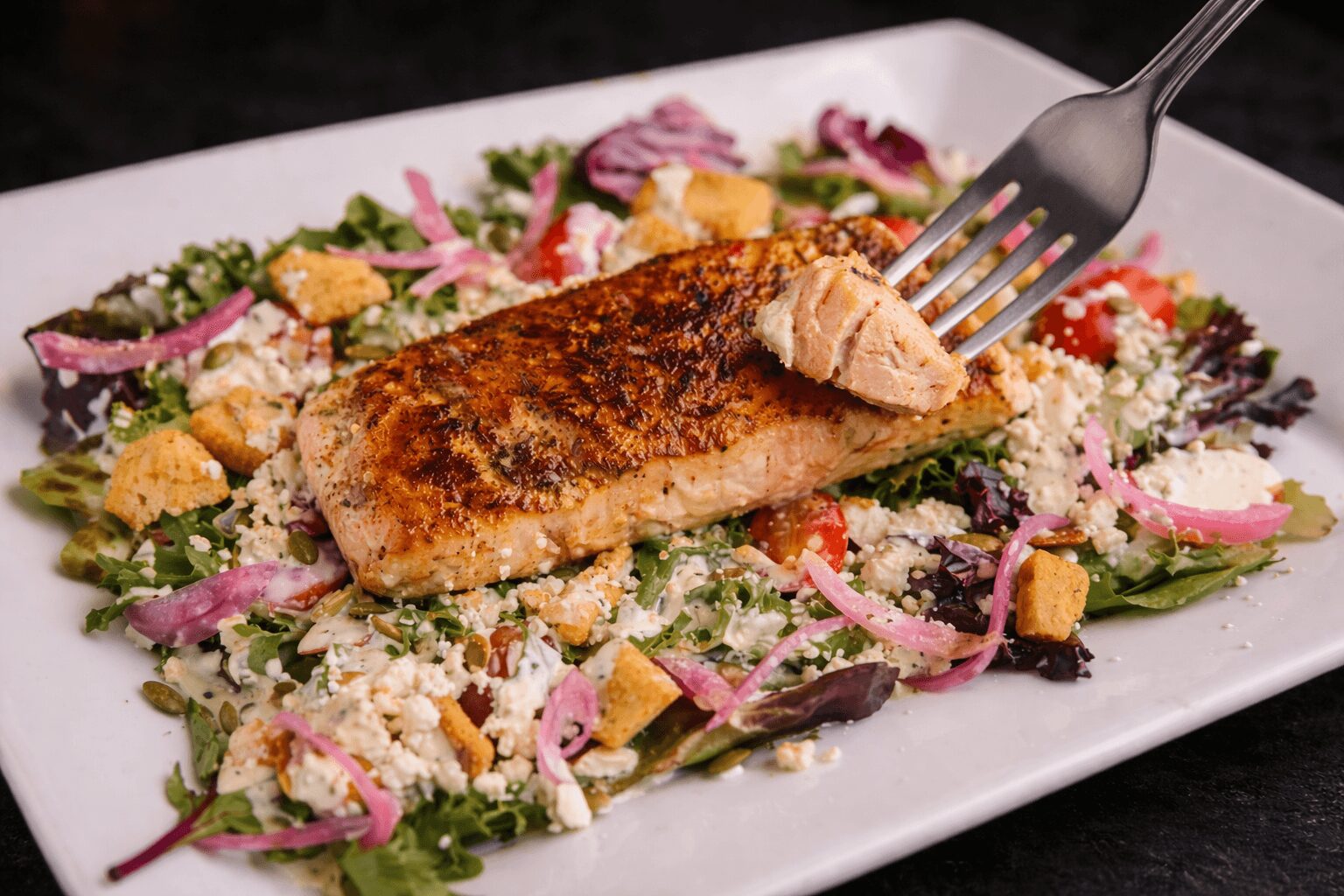 Grilled fish fillet on a bed of salad with vegetables and croutons on a white plate.