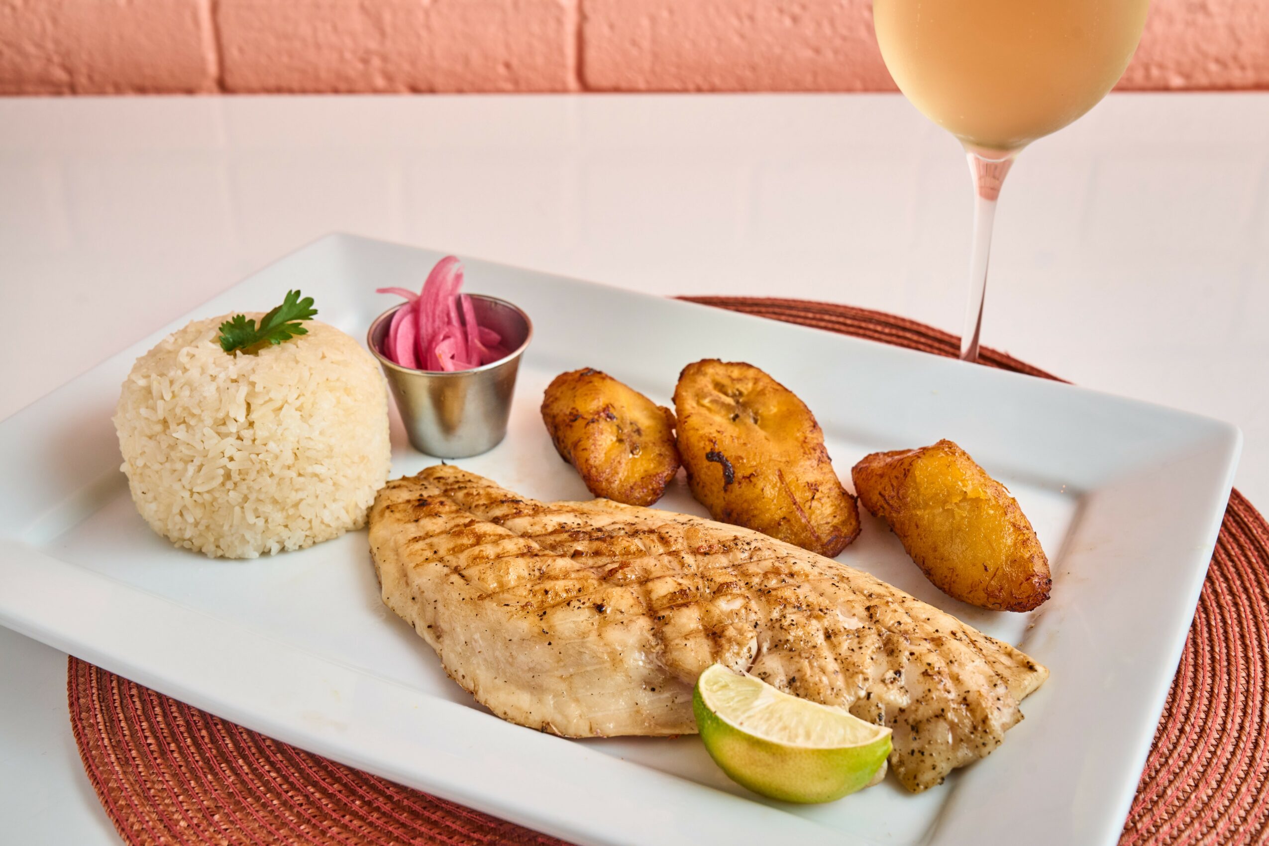 Grilled fish fillet with rice, fried plantains, pink pickled onions, lime wedge, and a drink in a glass, on a white plate.