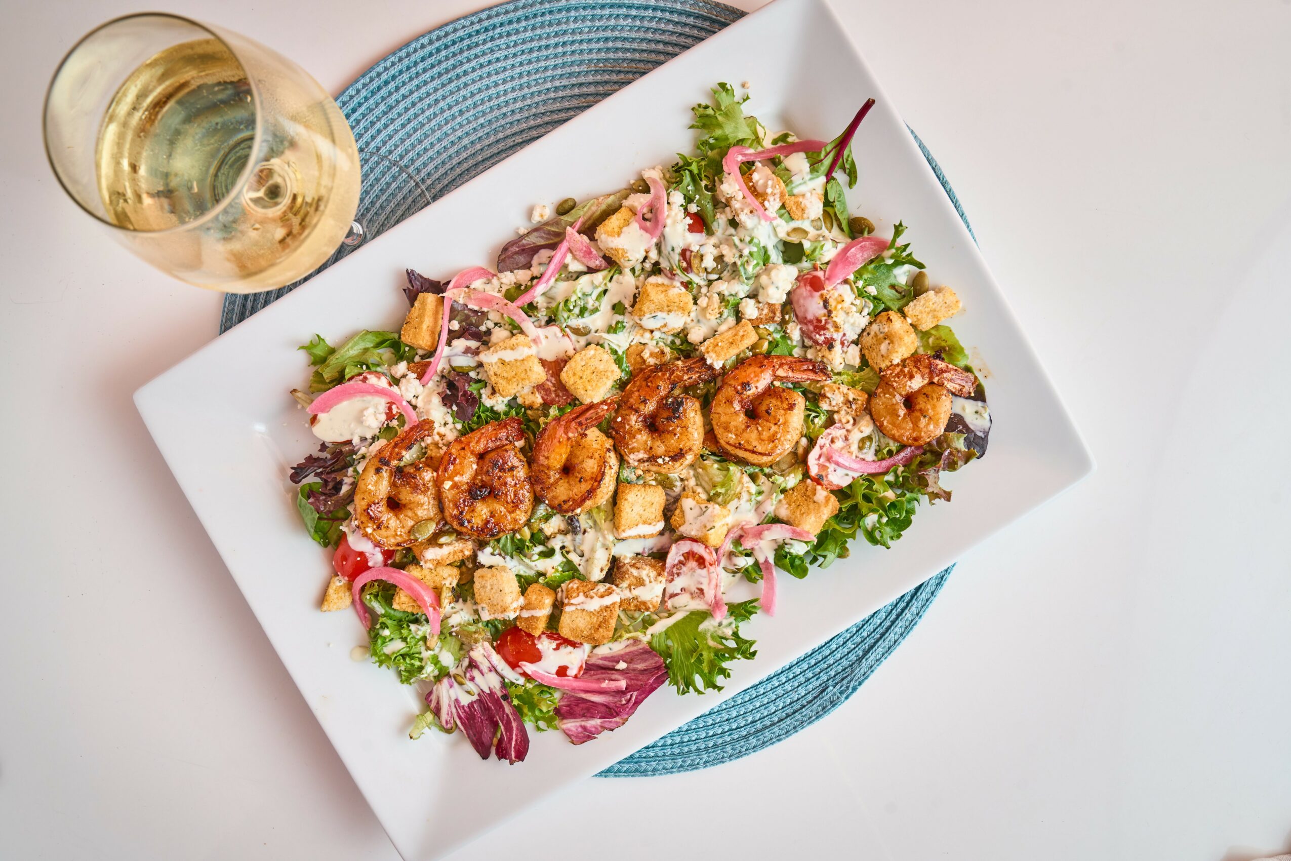 Salad with shrimp, vegetables, and dressing on a white plate, with a glass of white wine nearby.