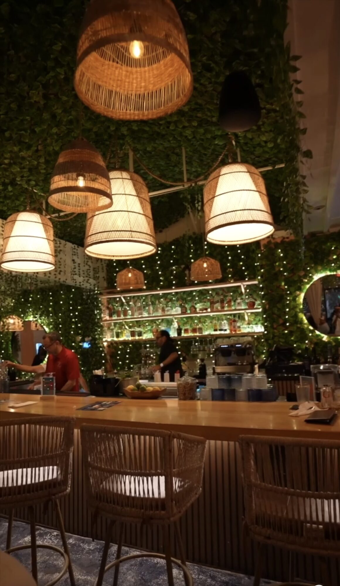 Bar with hanging woven lamps, shelves with bottles, and string lights, with people working behind the counter.