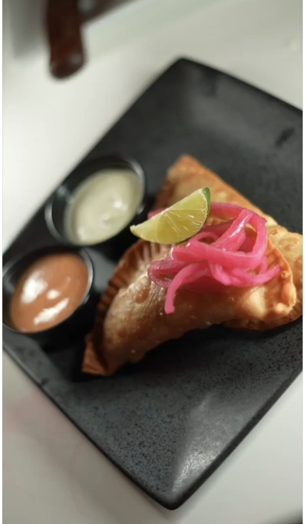 A slice of pizza with pink pickled onions and a lemon wedge on top, served with three dipping sauces on a black plate.