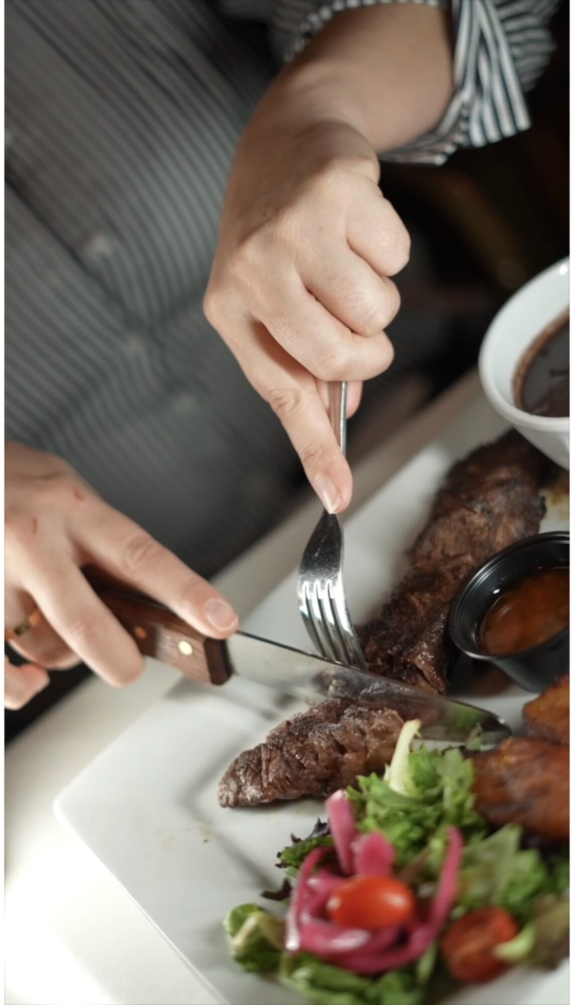 Person cutting cooked meat with fork and knife on a white plate, with salad and sauce nearby.
