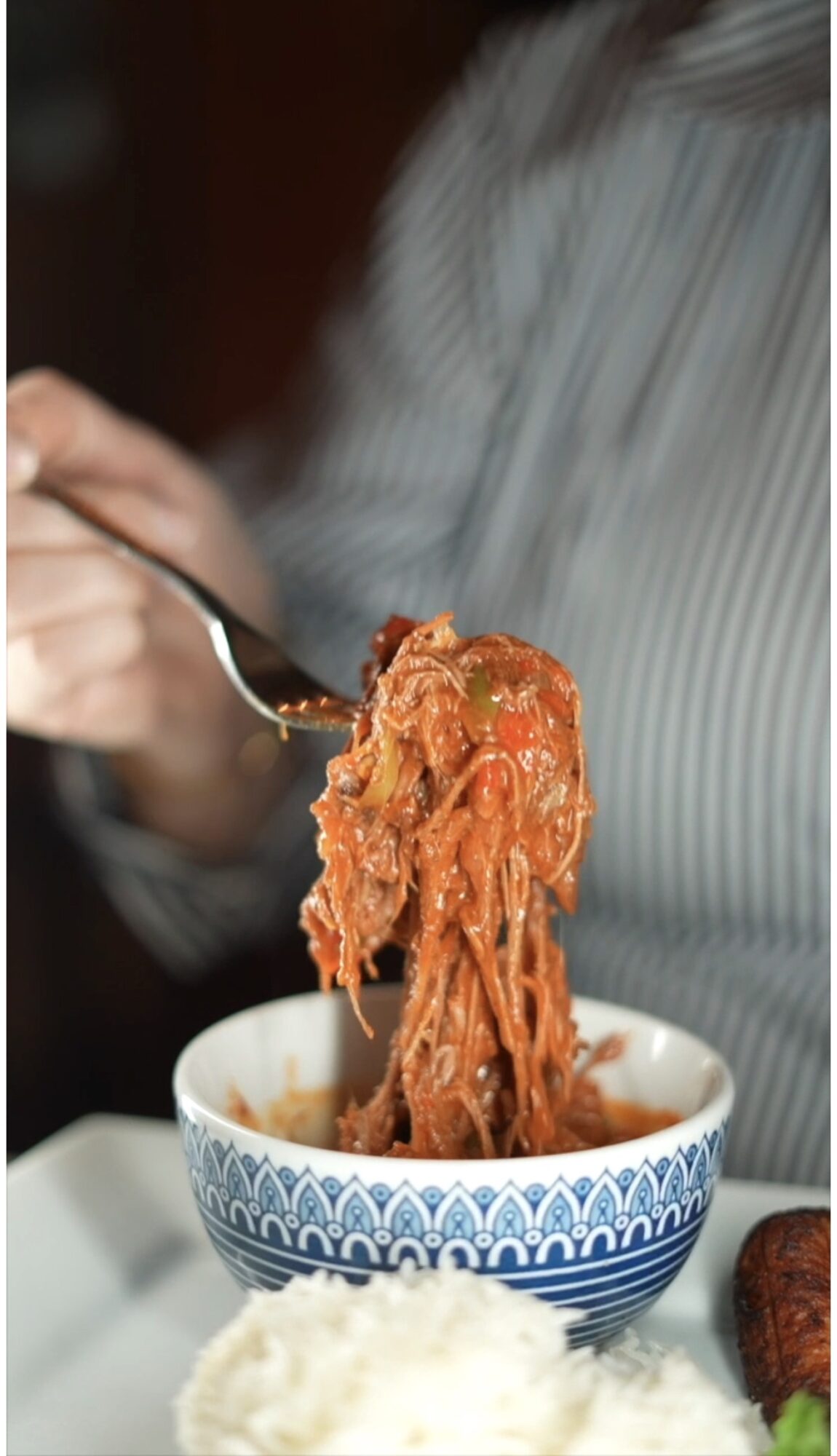 Person holding fork with spaghetti and tomato sauce over a patterned bowl, with rice on the side.