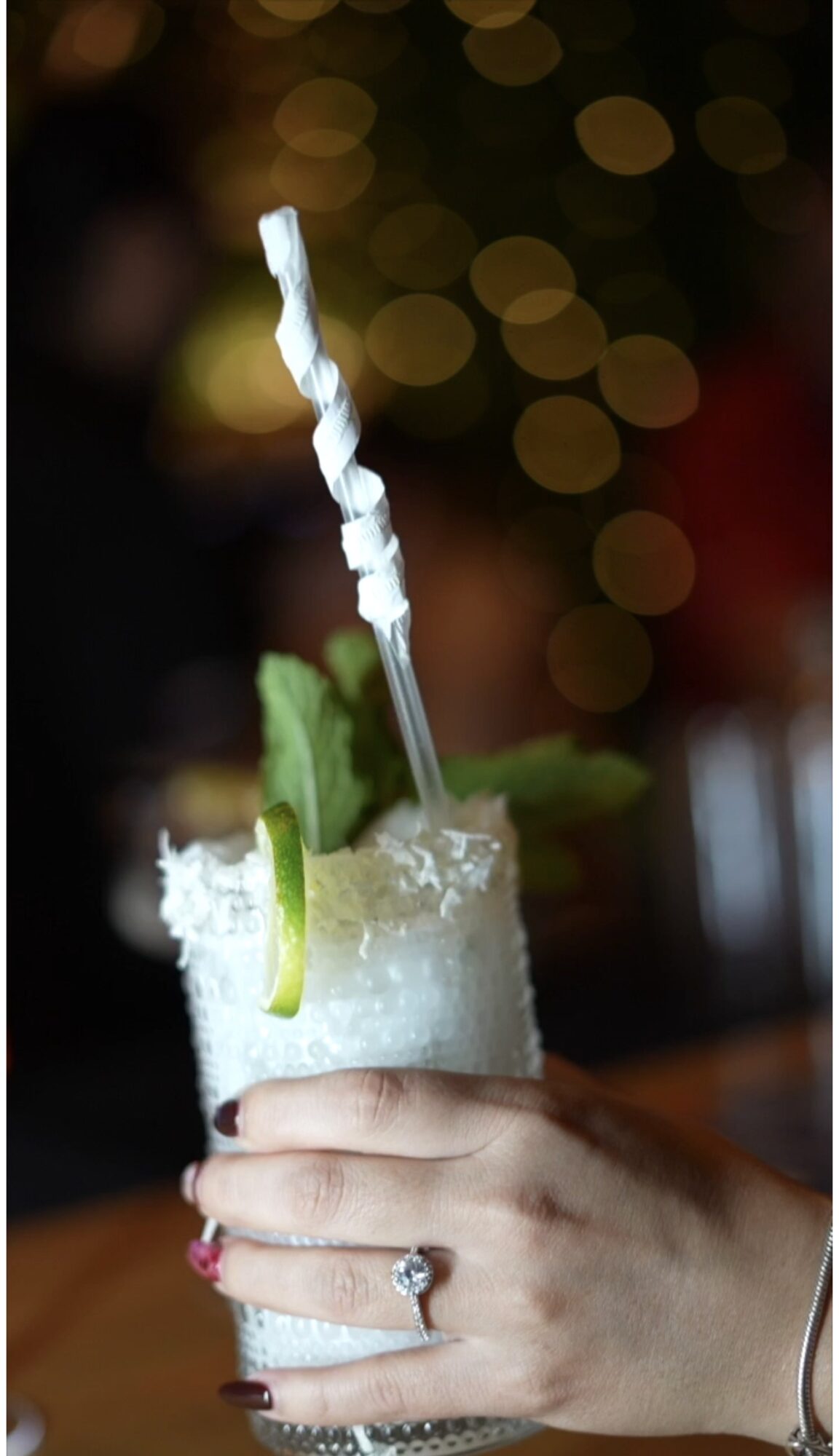 Hand holding a cocktail glass with lime, mint, straw, and sugar rim, against a bokeh background.