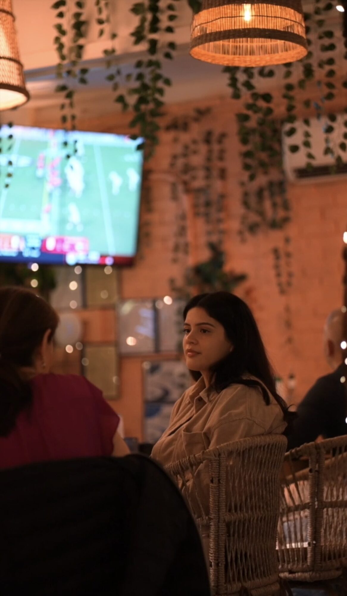 Young woman with long dark hair sitting at a table in a restaurant, engaging in conversation with others.