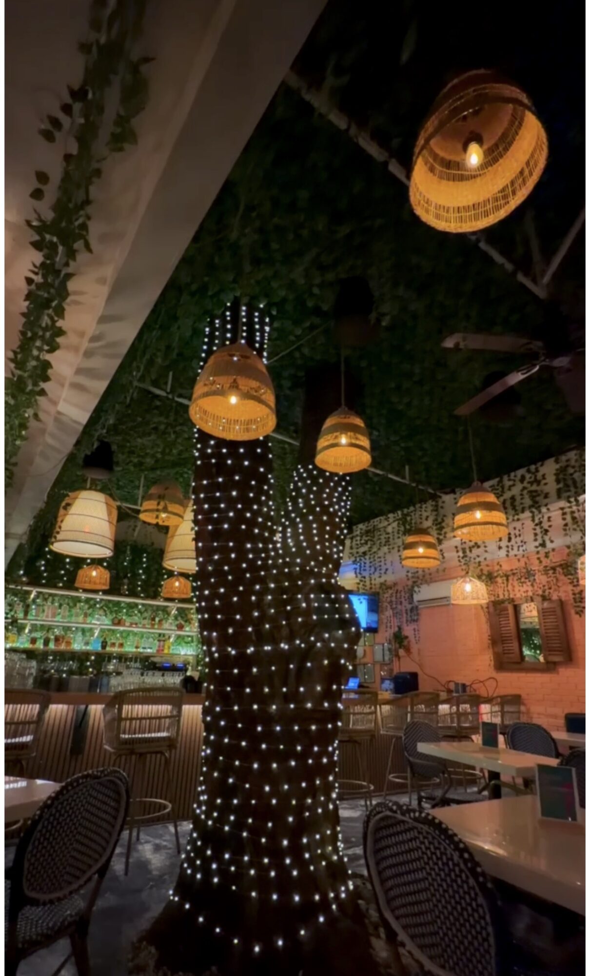 Indoor restaurant with hanging lamps and a decorated tree with lights, tables, and chairs visible.