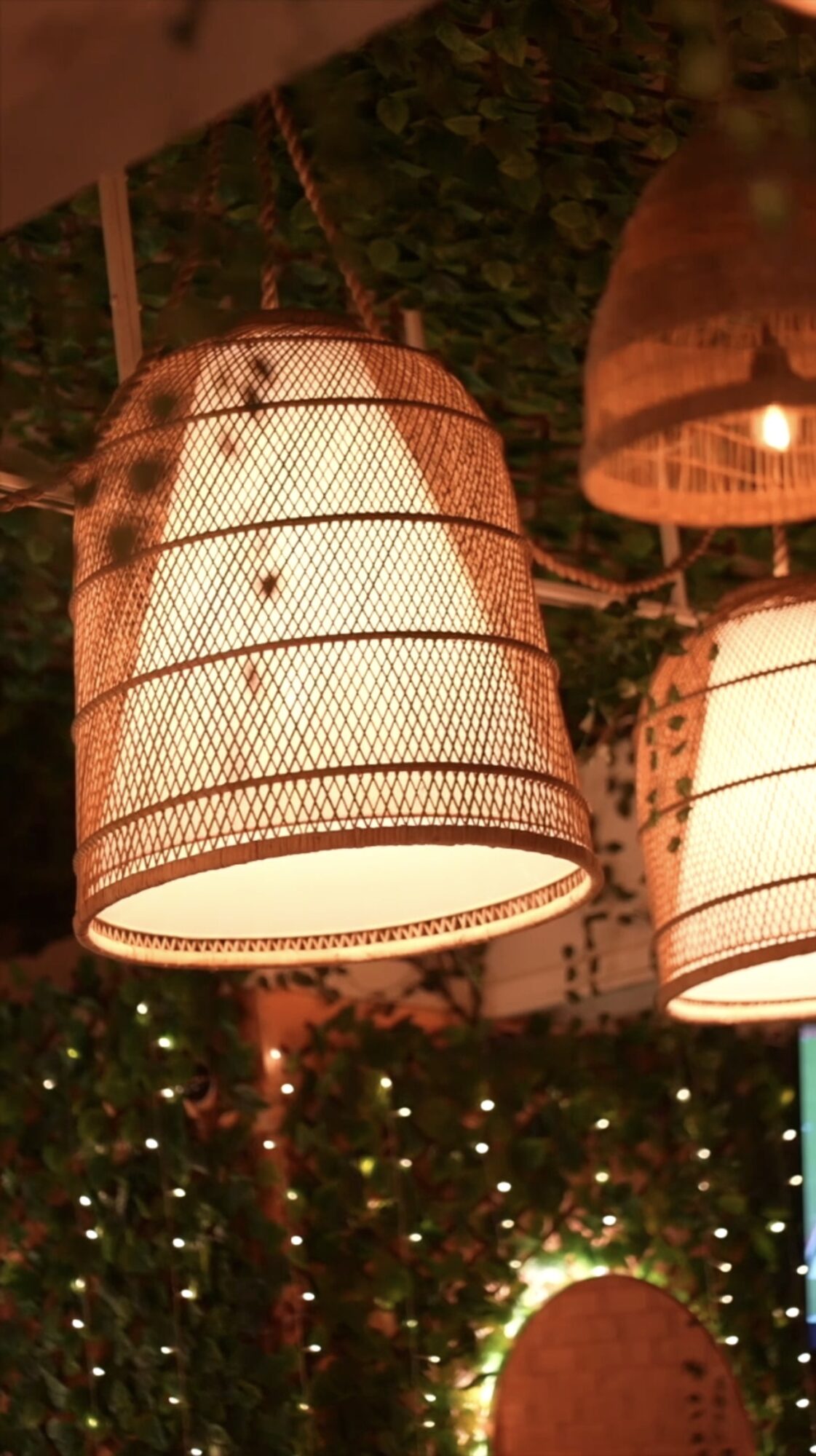 Two hanging woven lamps illuminated, with greenery and string lights in the background.