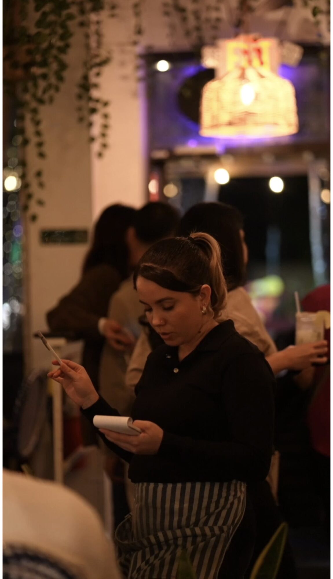Woman with striped pants and dark top looking at her phone in a dimly lit indoor setting.