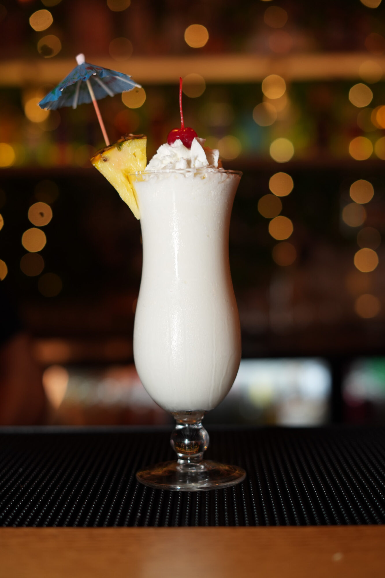 Tall glass with a creamy white drink, garnished with a cherry, pineapple slice, and a small blue umbrella.