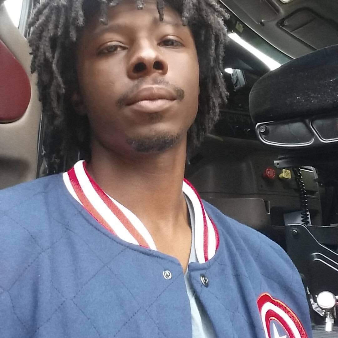 Young man with dreadlocks wearing a blue jacket with red and white collar, sitting inside a vehicle.