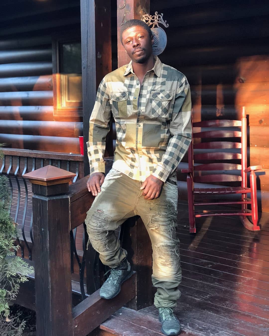 Man standing on porch with wooden railing and chair, wearing a checkered shirt and jeans, outdoors during daytime.