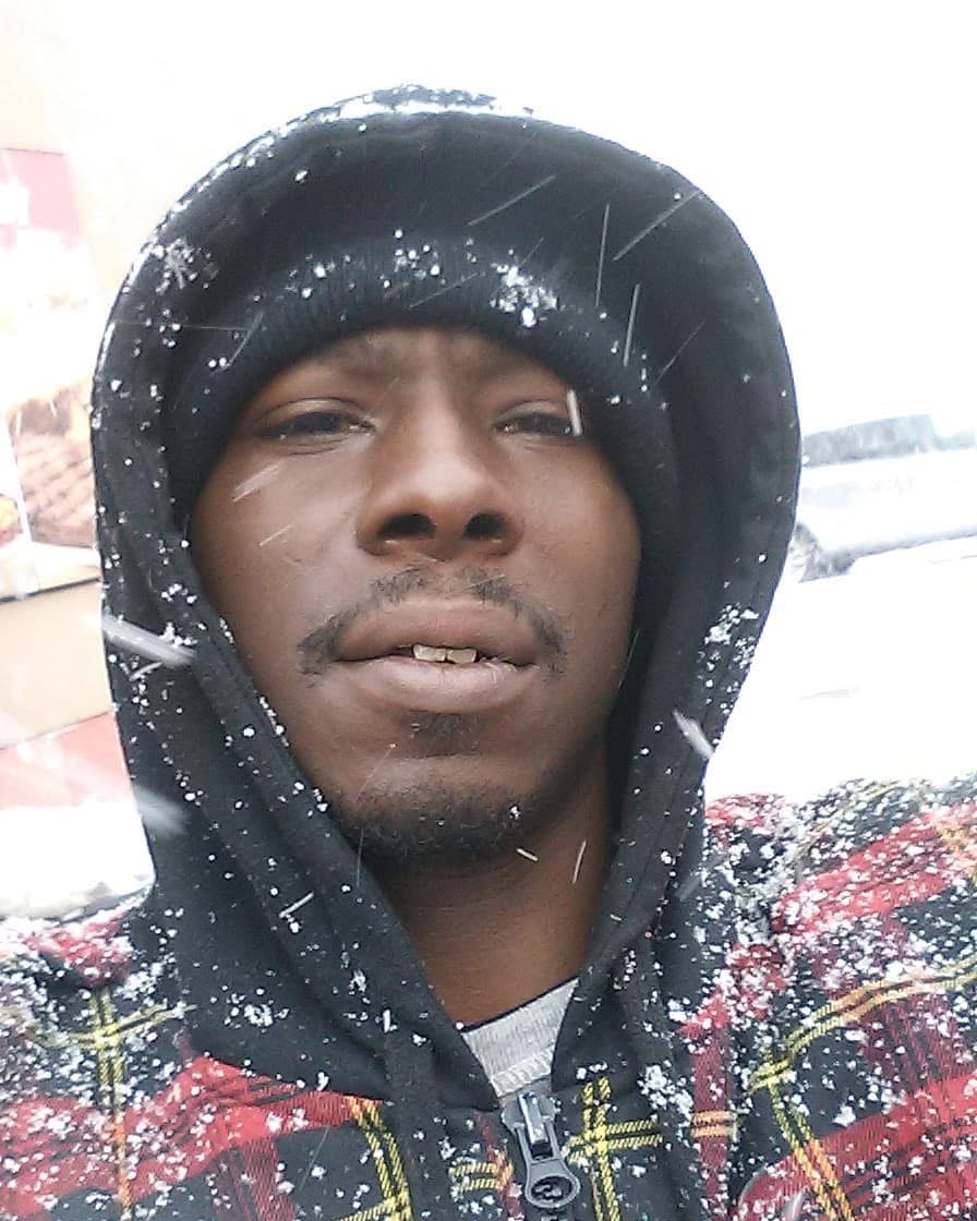 Person wearing a black hoodie with snow on it, outdoors in snowy weather, looking at the camera.