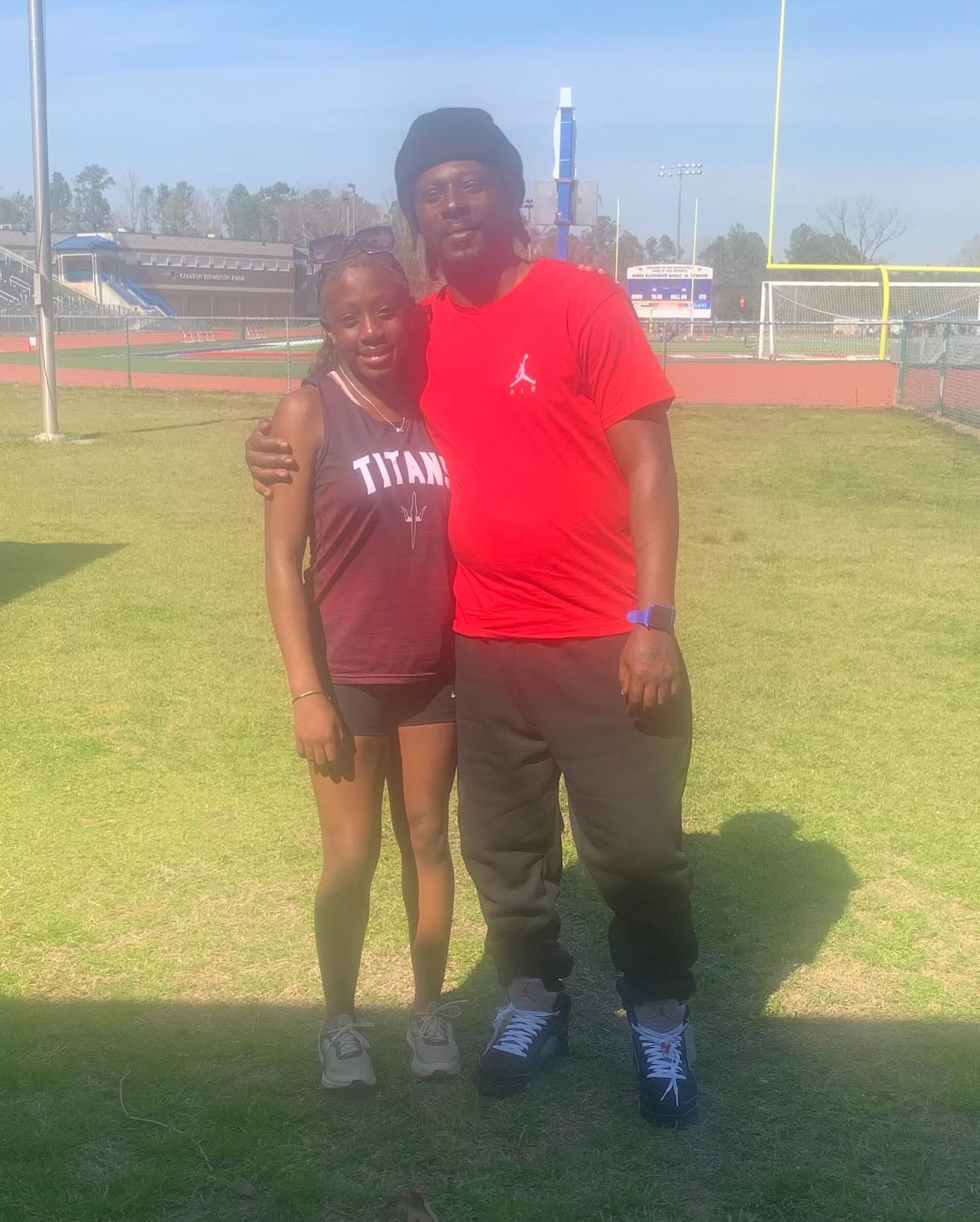 Two people standing on a grassy field, one with arm around the other, both smiling, sunny day, sports field background.