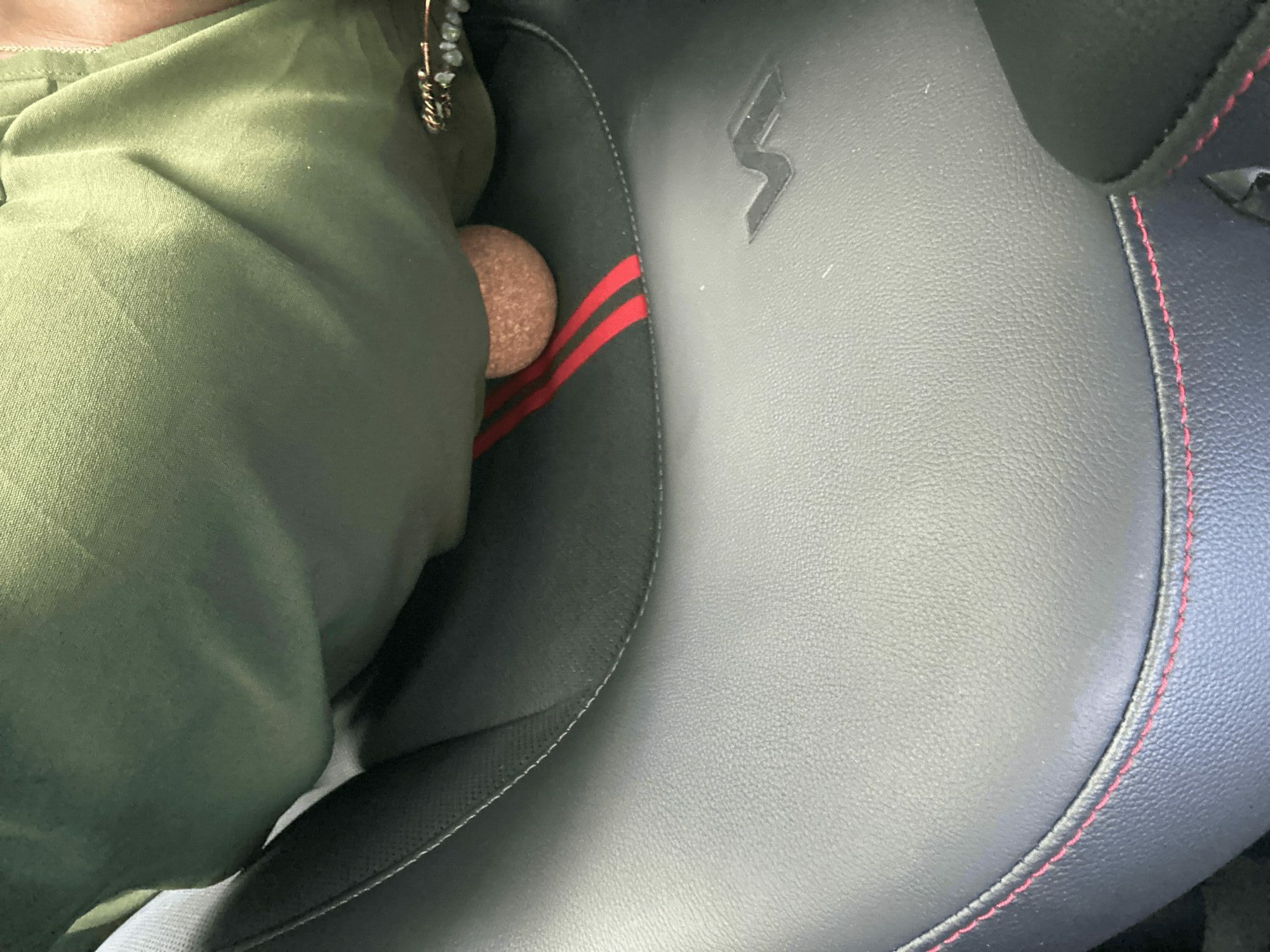 Part of a person's leg in green pants and a shoe, sitting on a black seat with red stitching.