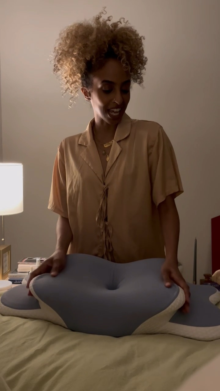 Woman with curly hair in a beige shirt, smiling, touching a gray cushion on bed, in a softly lit room.