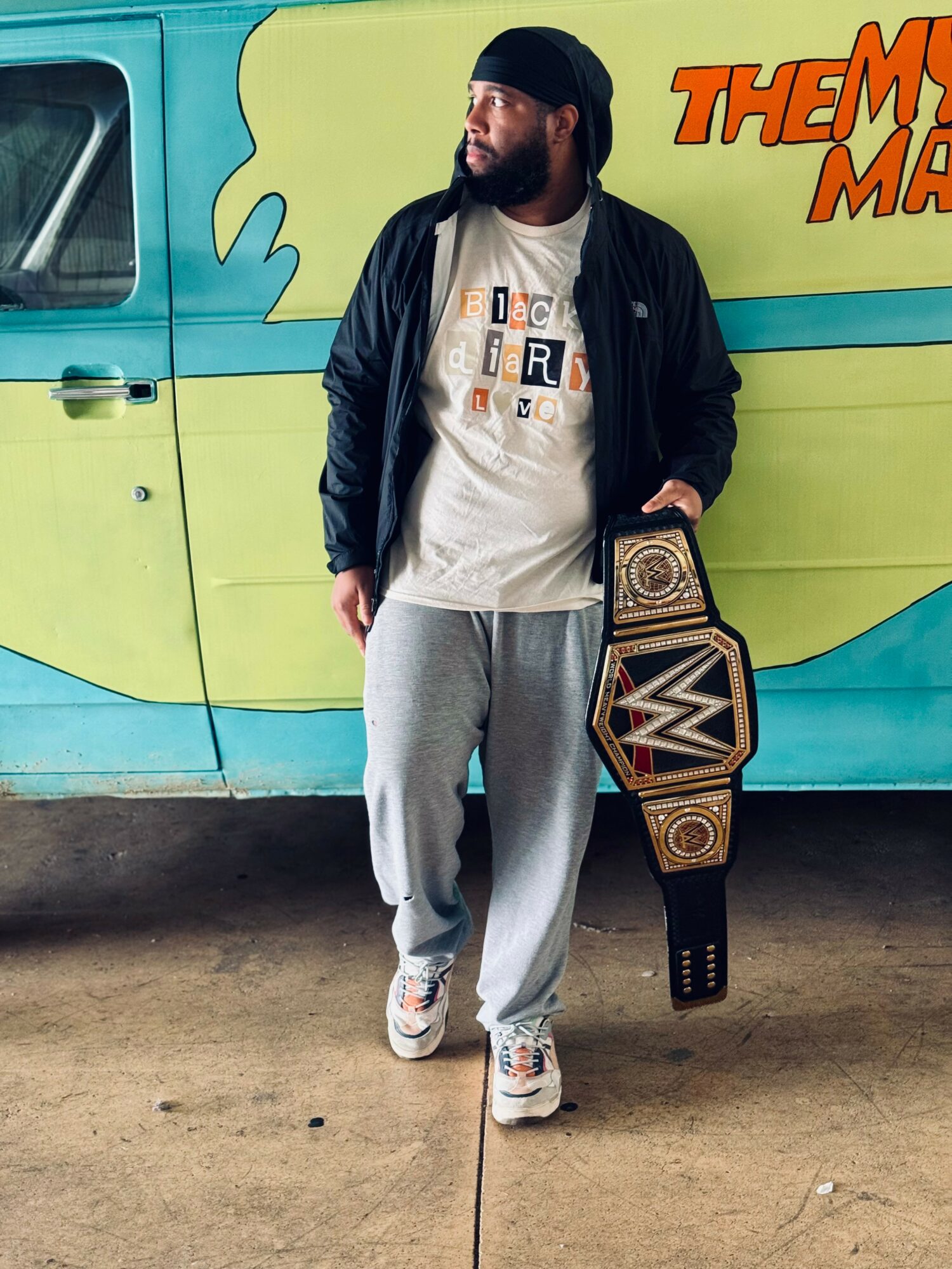 Man with long hair and beard holding championship belt, standing in front of colorful van with text, wearing casual clothes.