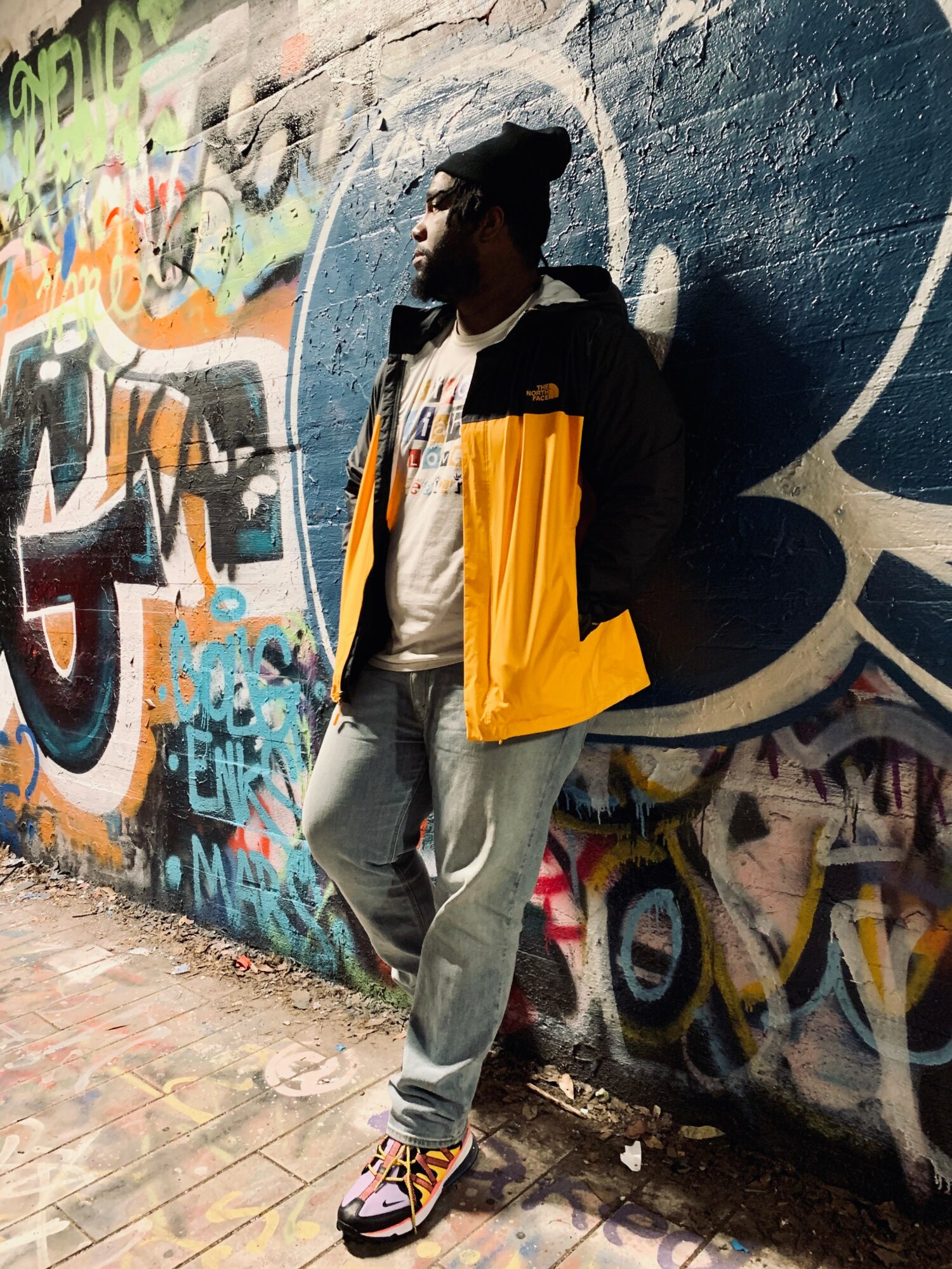 Person leaning against a graffiti-covered wall wearing a yellow and black jacket, gray pants, and sneakers.