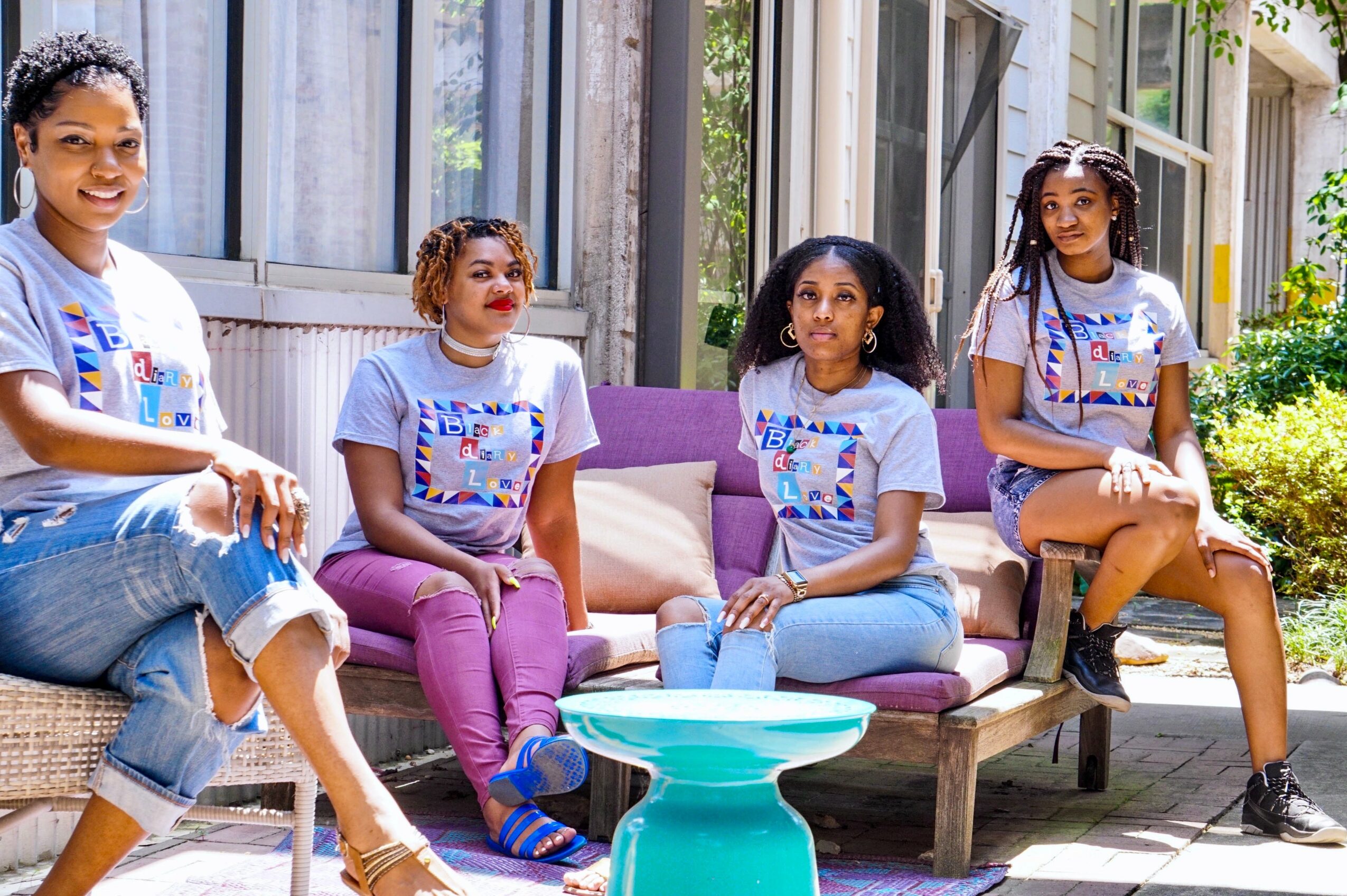 Four girls sitting outdoors on a purple sofa and a chair, smiling, with a building and greenery behind them.