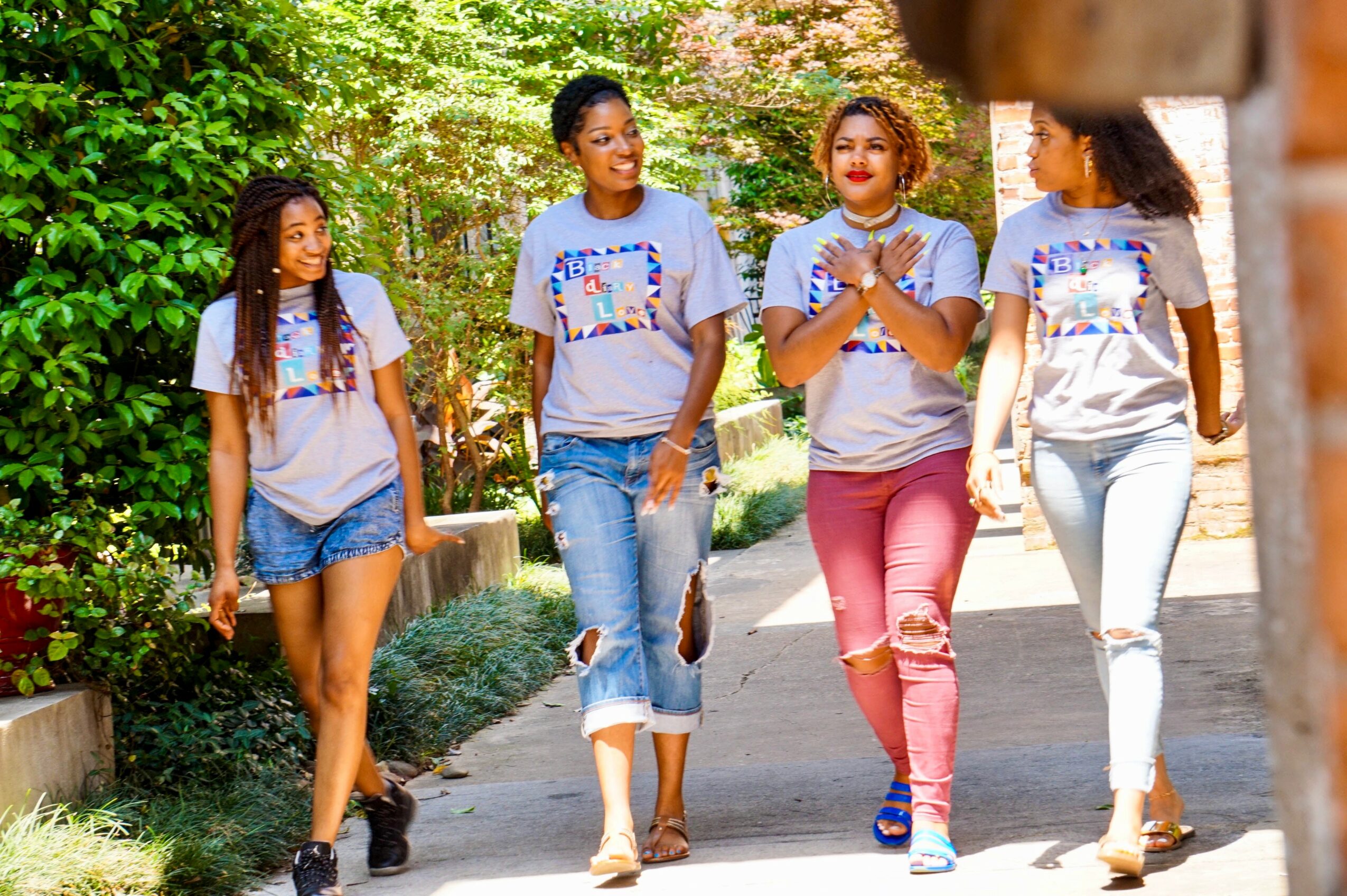 Four women walking outdoors on a sunny day, surrounded by greenery, smiling and talking.
