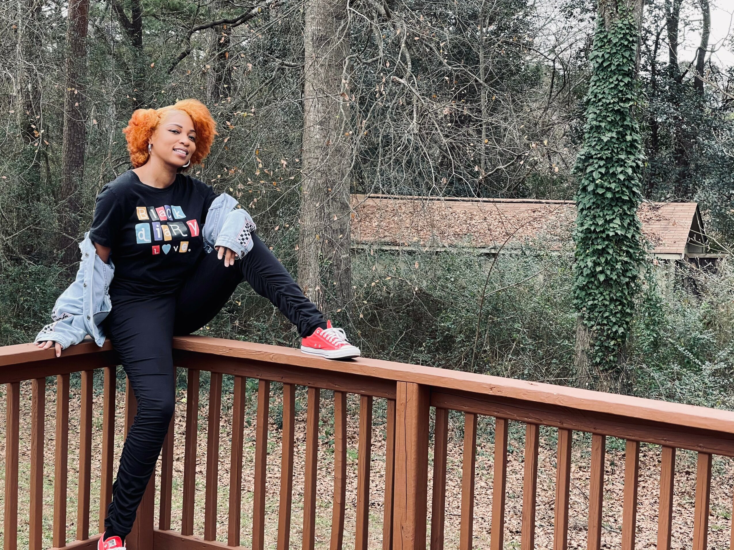 Person with orange hair leaning on a wooden railing outdoors, trees and a building in the background.