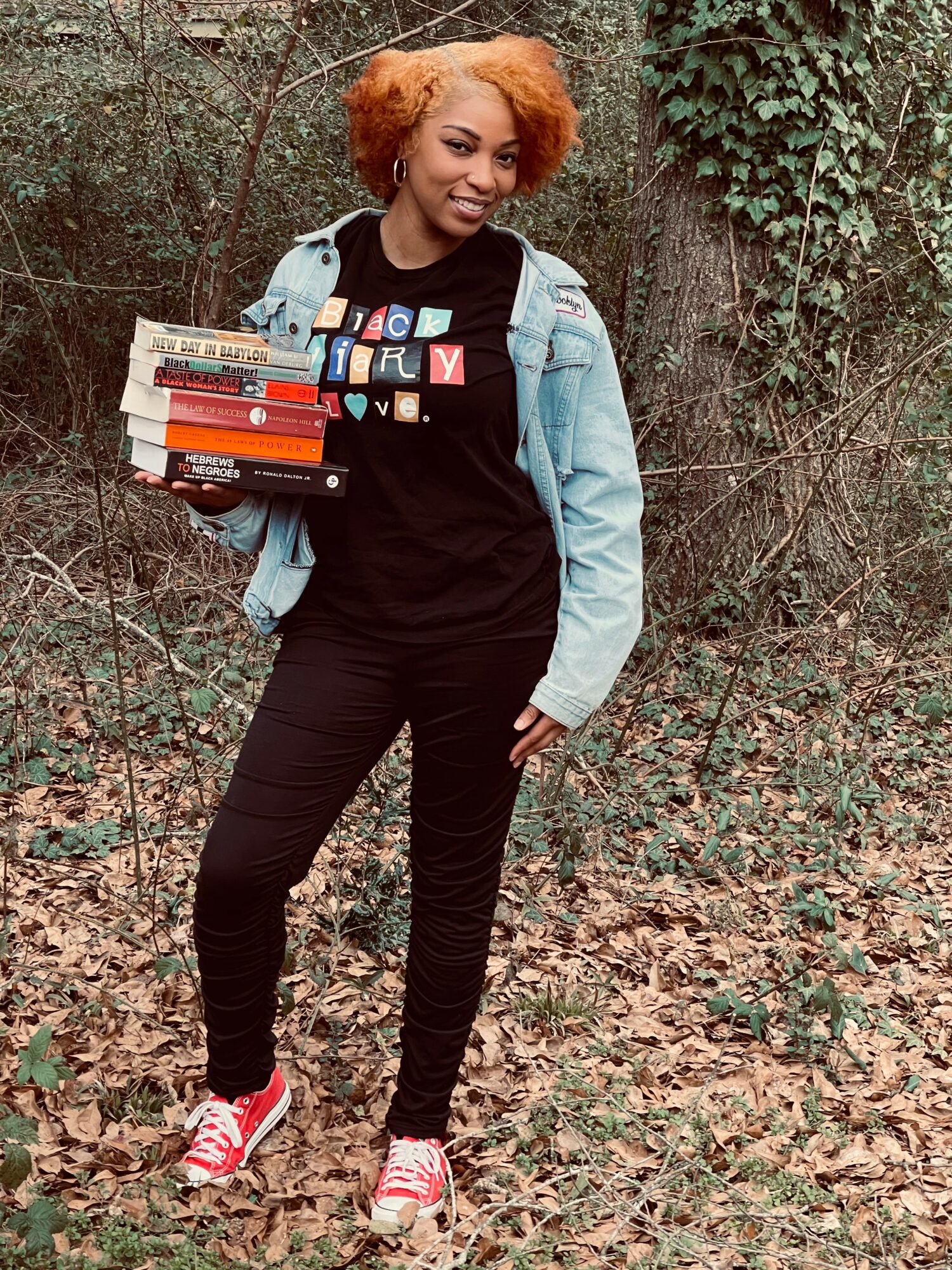 Young woman with orange hair holding a stack of books outdoors, wearing a black shirt, denim jacket, black pants, and red sneakers.