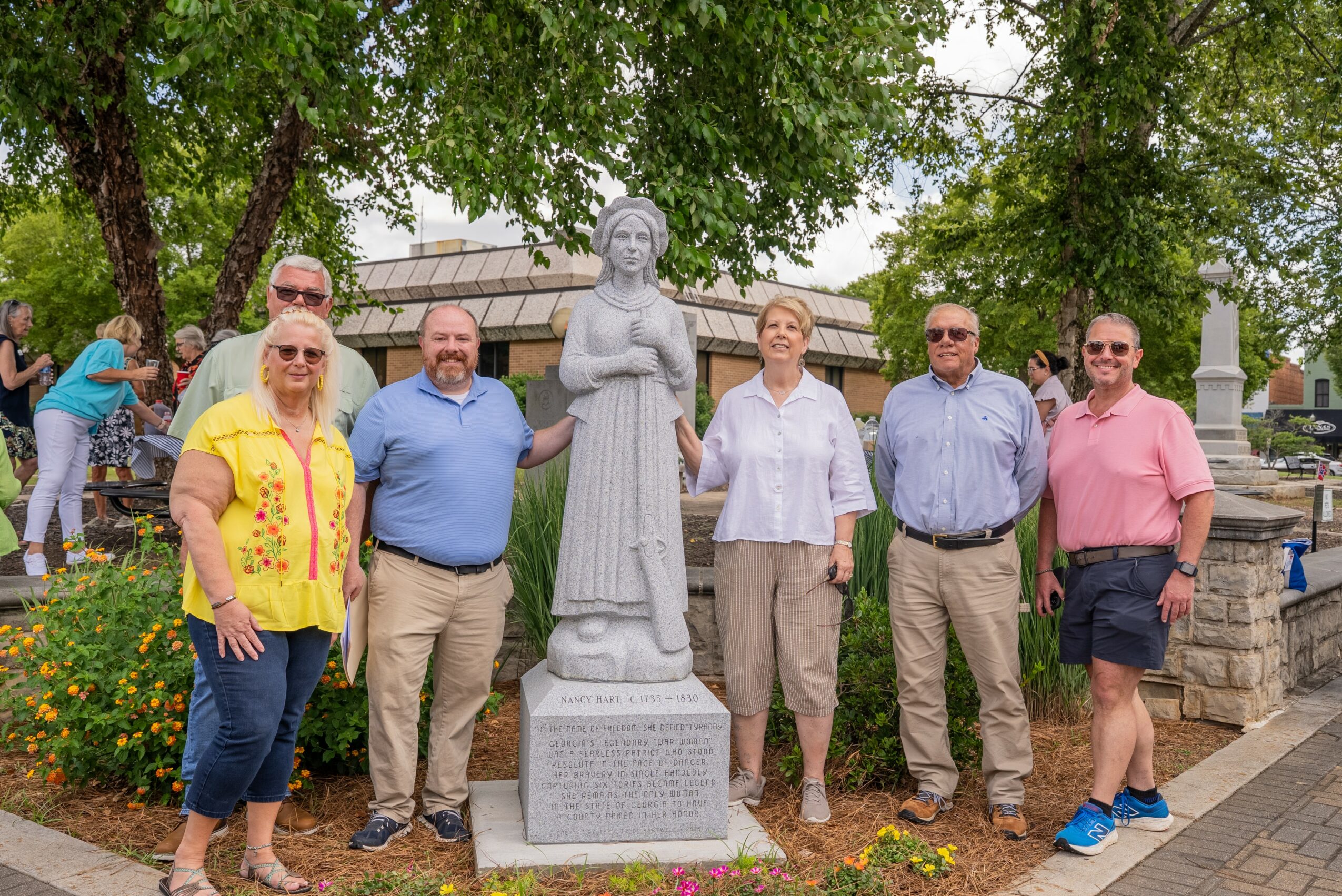 Six people stand in front of a statue in a park with trees and flowers, smiling at the camera.