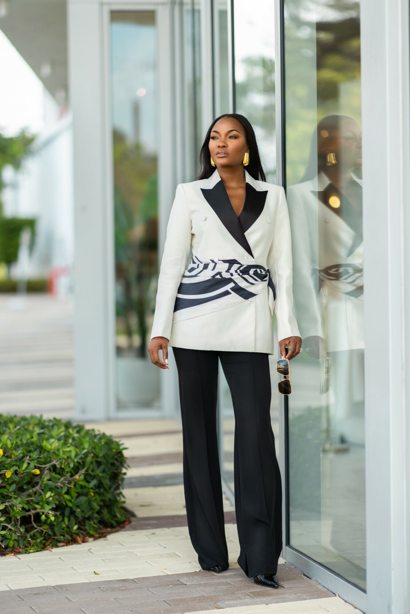 Woman in white blazer and black pants standing outside near glass building, holding sunglasses, with greenery nearby.
