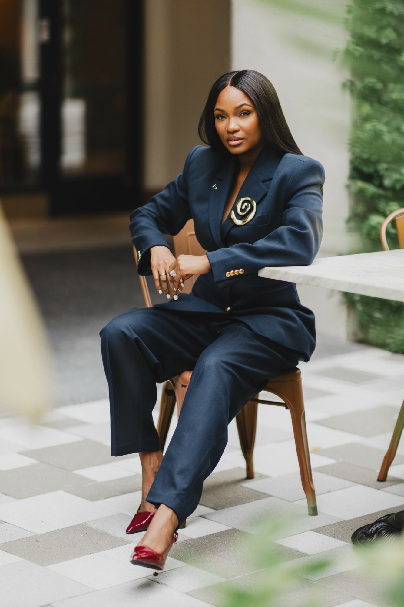 Woman in navy suit sitting on a chair outdoors, with greenery and a white table nearby.