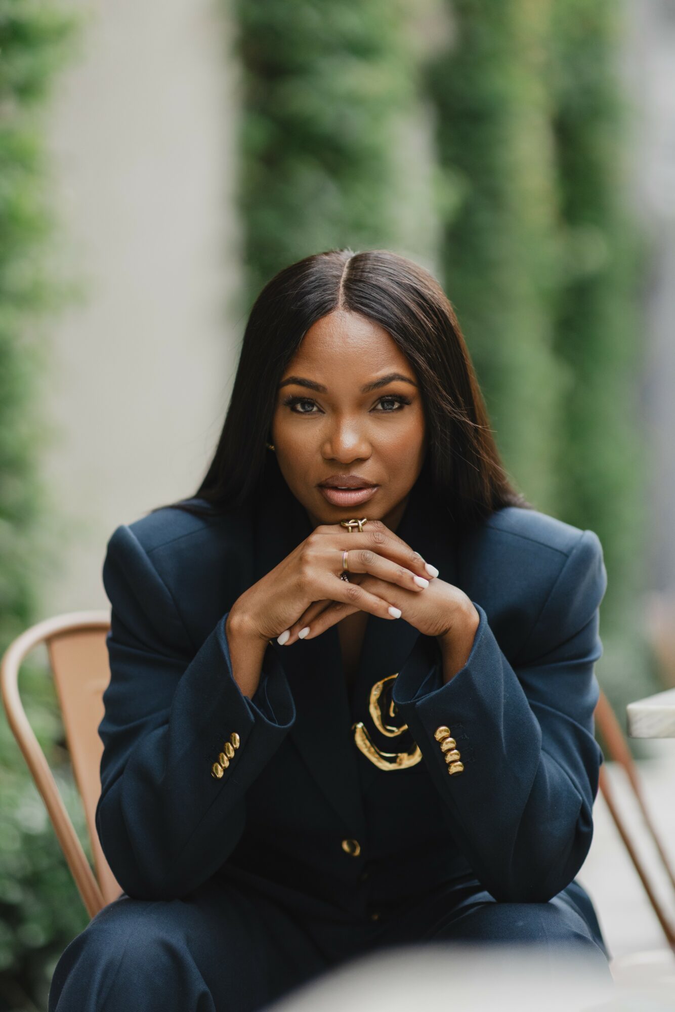 Woman with long dark hair in a navy blazer sitting outdoors with greenery in background.