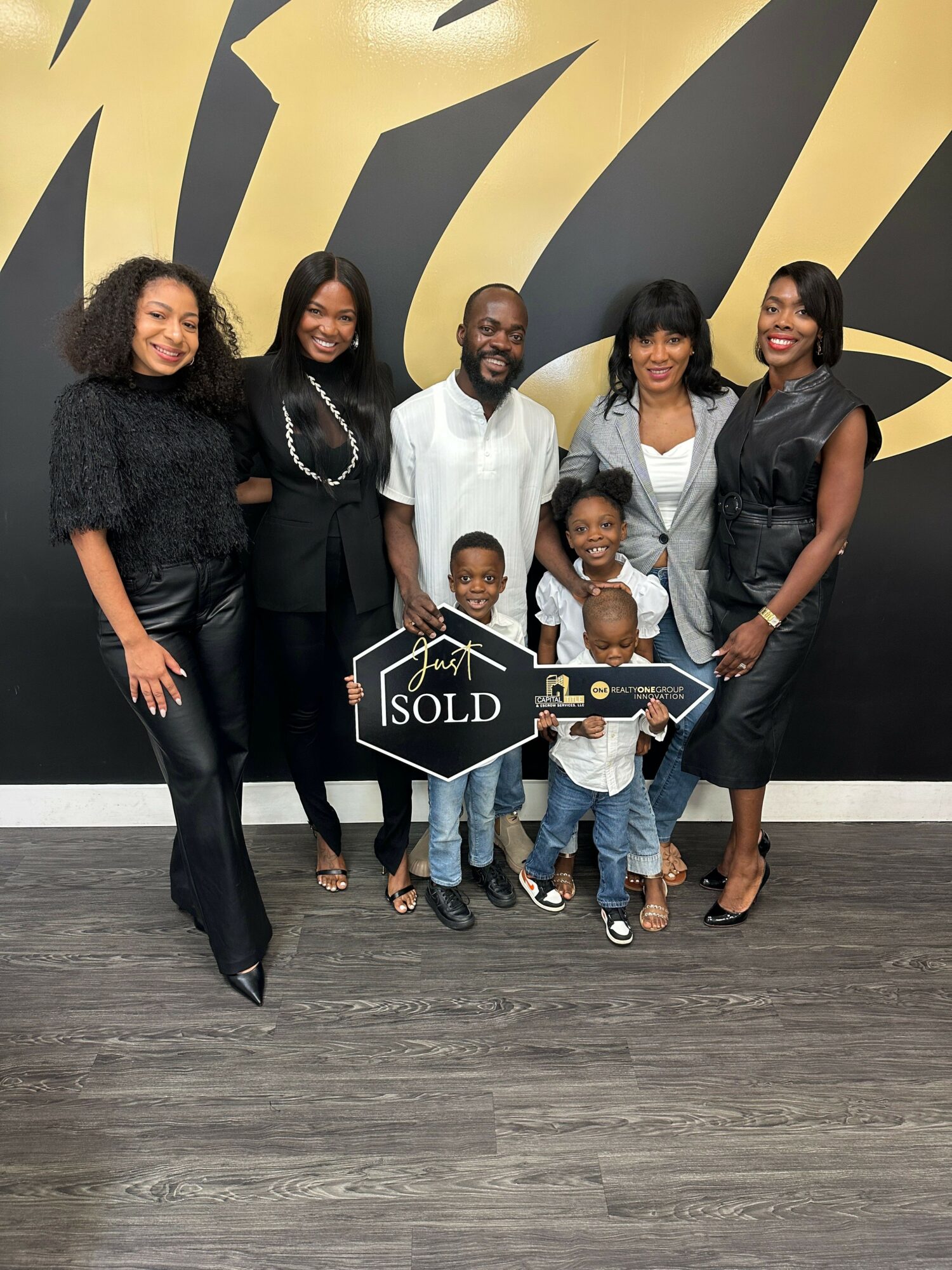 Group of six people, including children, standing indoors with a large black and gold abstract background, holding a house-shaped 'SOLD' sign.