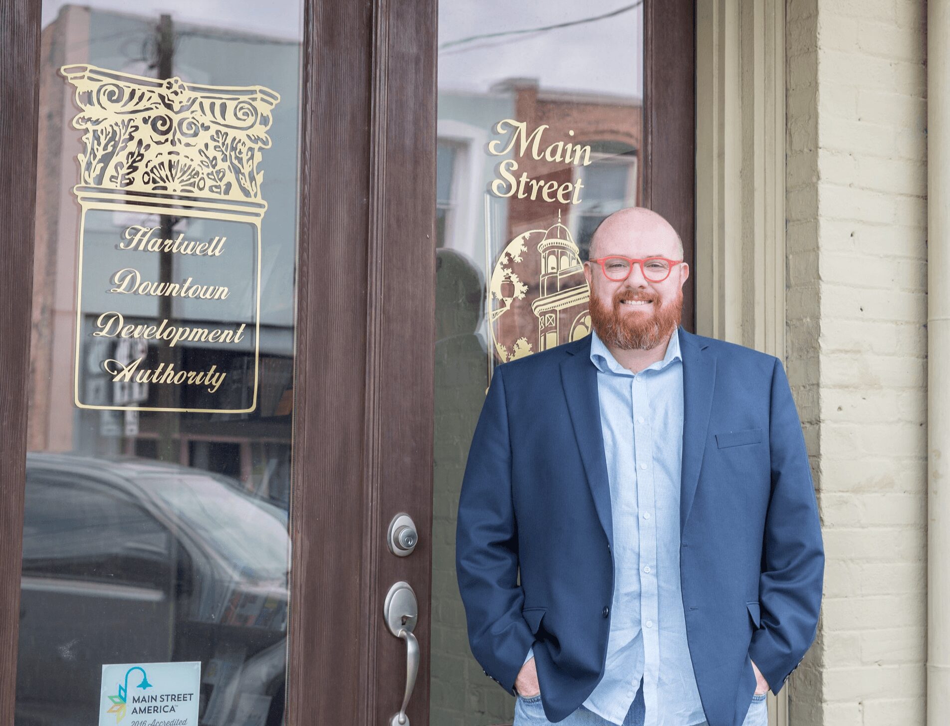Man with glasses, beard, in a blue blazer and light shirt, standing outside a building with glass door and window.