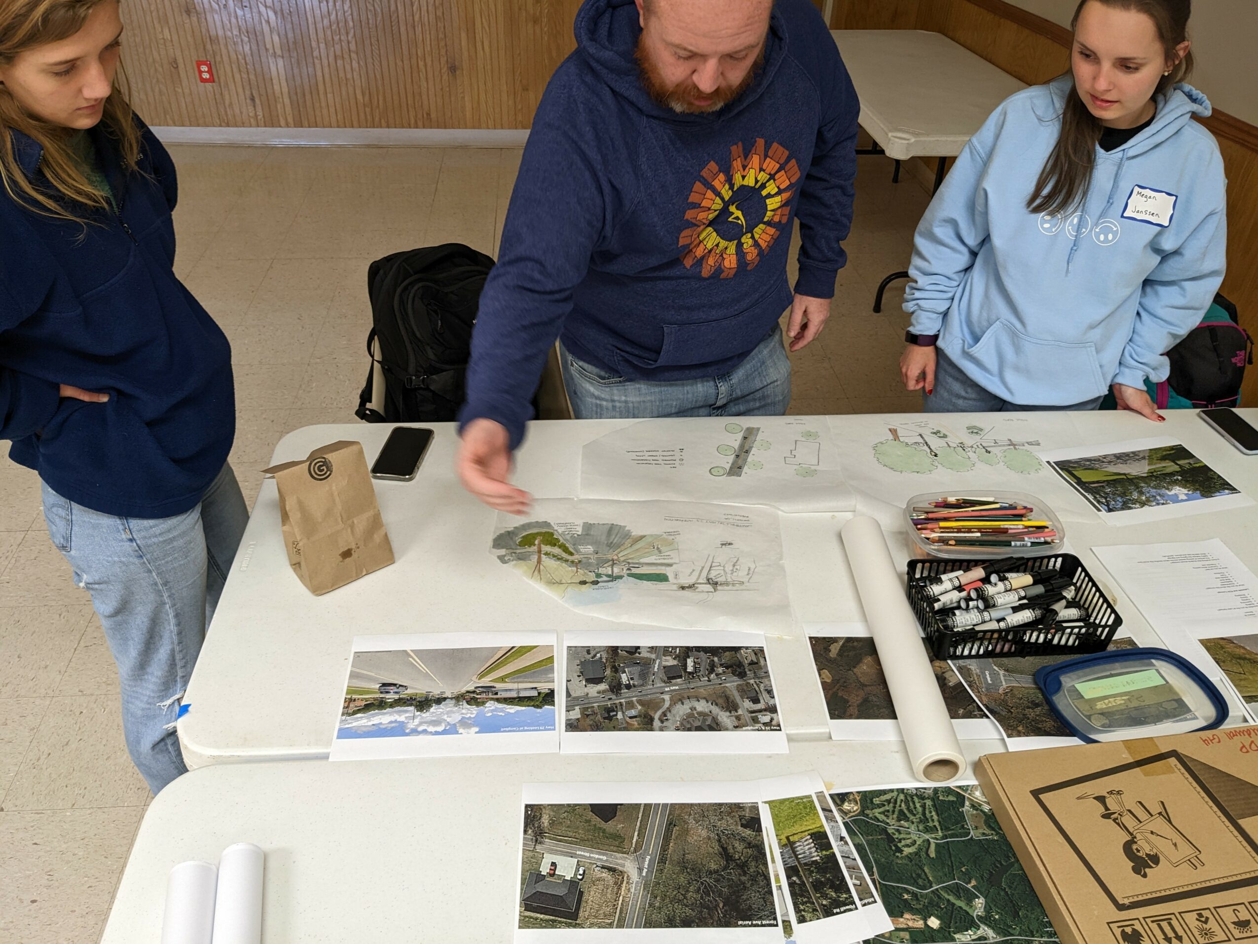 Three people around a table with photos, papers, and a cardboard box, indoors with wooden walls.