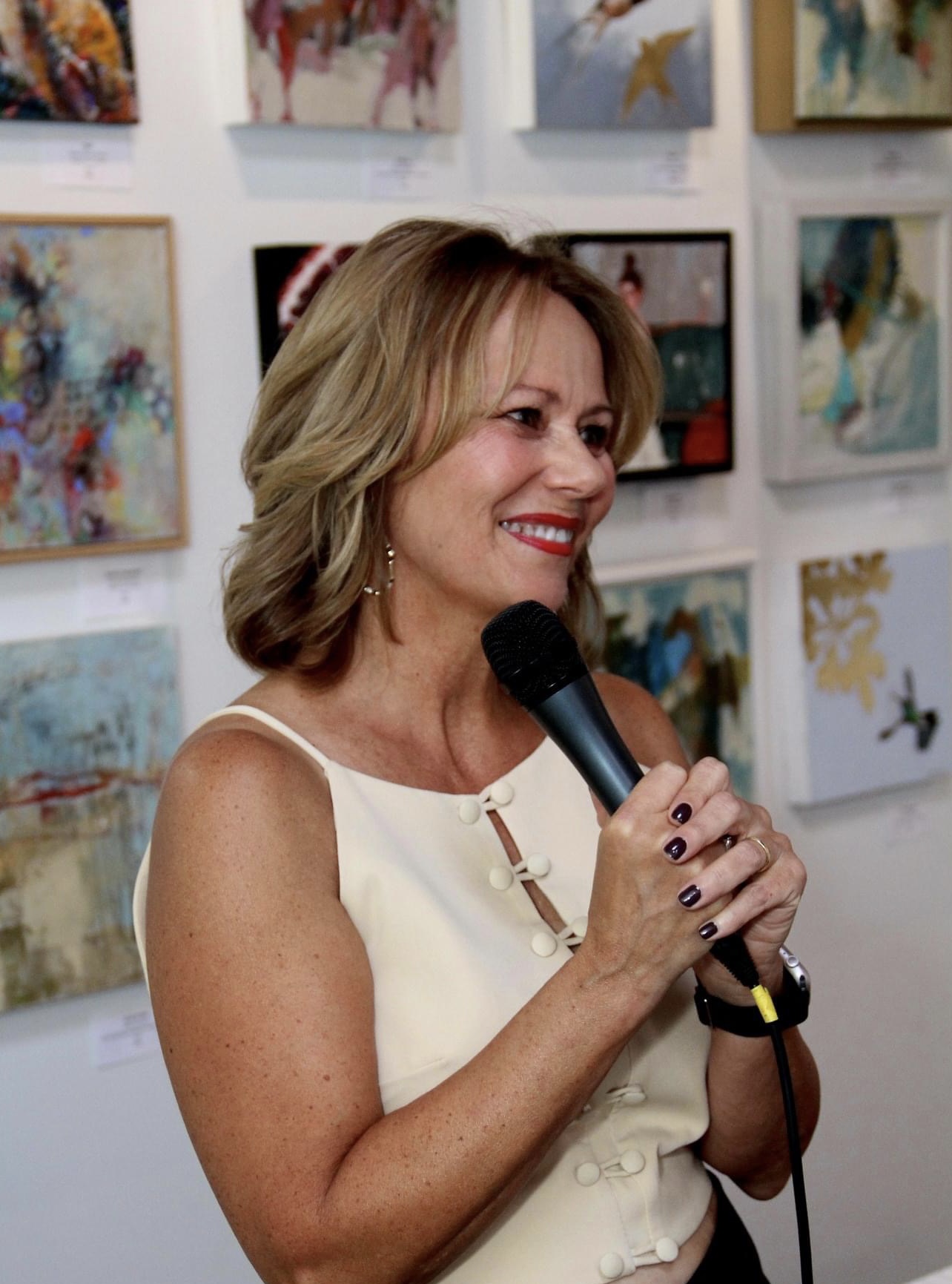 Woman with shoulder-length hair smiling, holding a microphone, in front of colorful artwork on gallery wall.