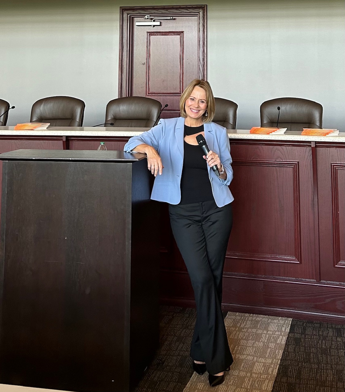 Woman in blue blazer holding microphone, standing in front of a panel with chairs and a door behind her.