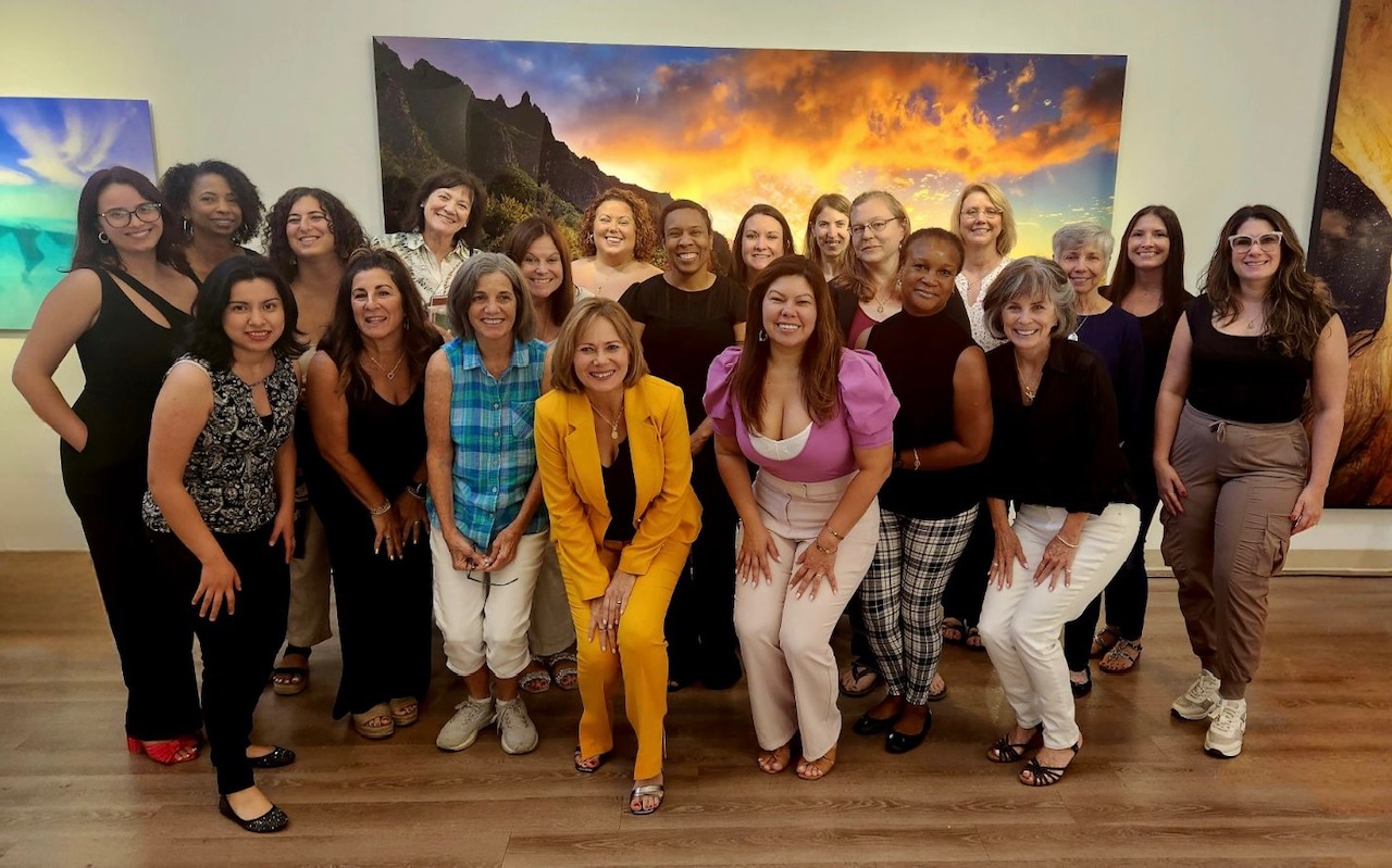 Group of 20 women standing indoors in front of a landscape painting, smiling and posing for a photo.